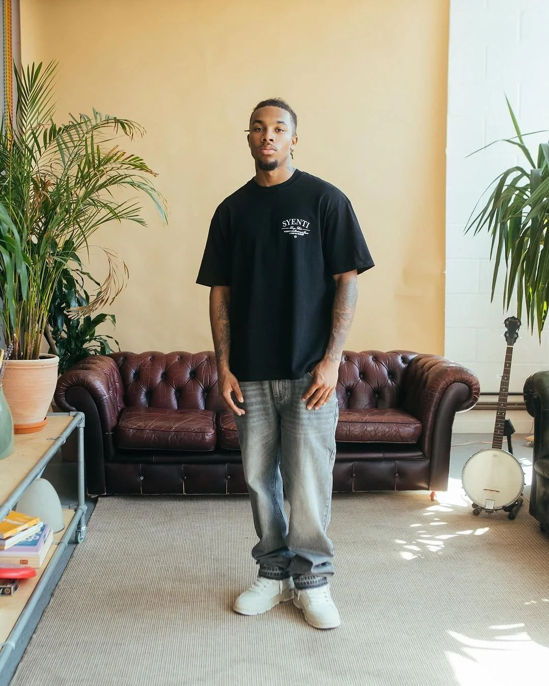 A young man standing in a room with beige walls, a brown leather sofa, large potted plants, a banjo, and a table with books and decorative items.