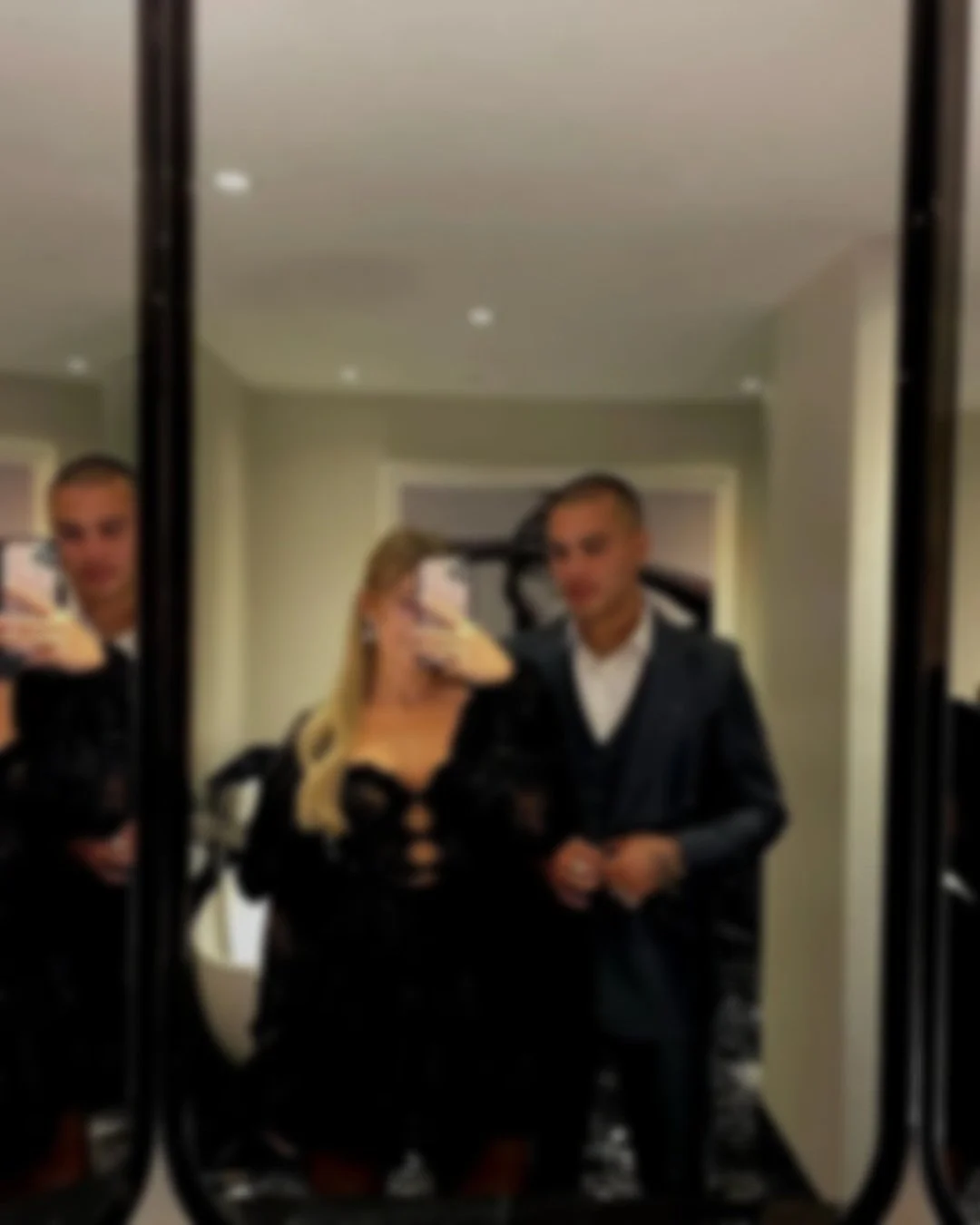 Three people taking a mirror selfie, with a woman in the center and two men on either side, in an indoor setting with a beige wall and door in the background. The image is blurry.