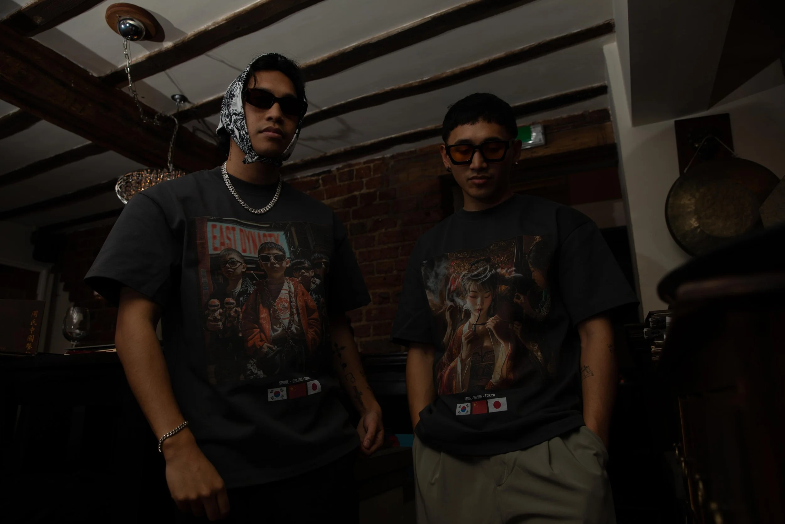 Two young men standing indoors, both wearing dark graphic t-shirts with Asian-themed images and flags, wearing sunglasses, with tattoos on their arms, in a room with exposed brick walls and wooden beams.