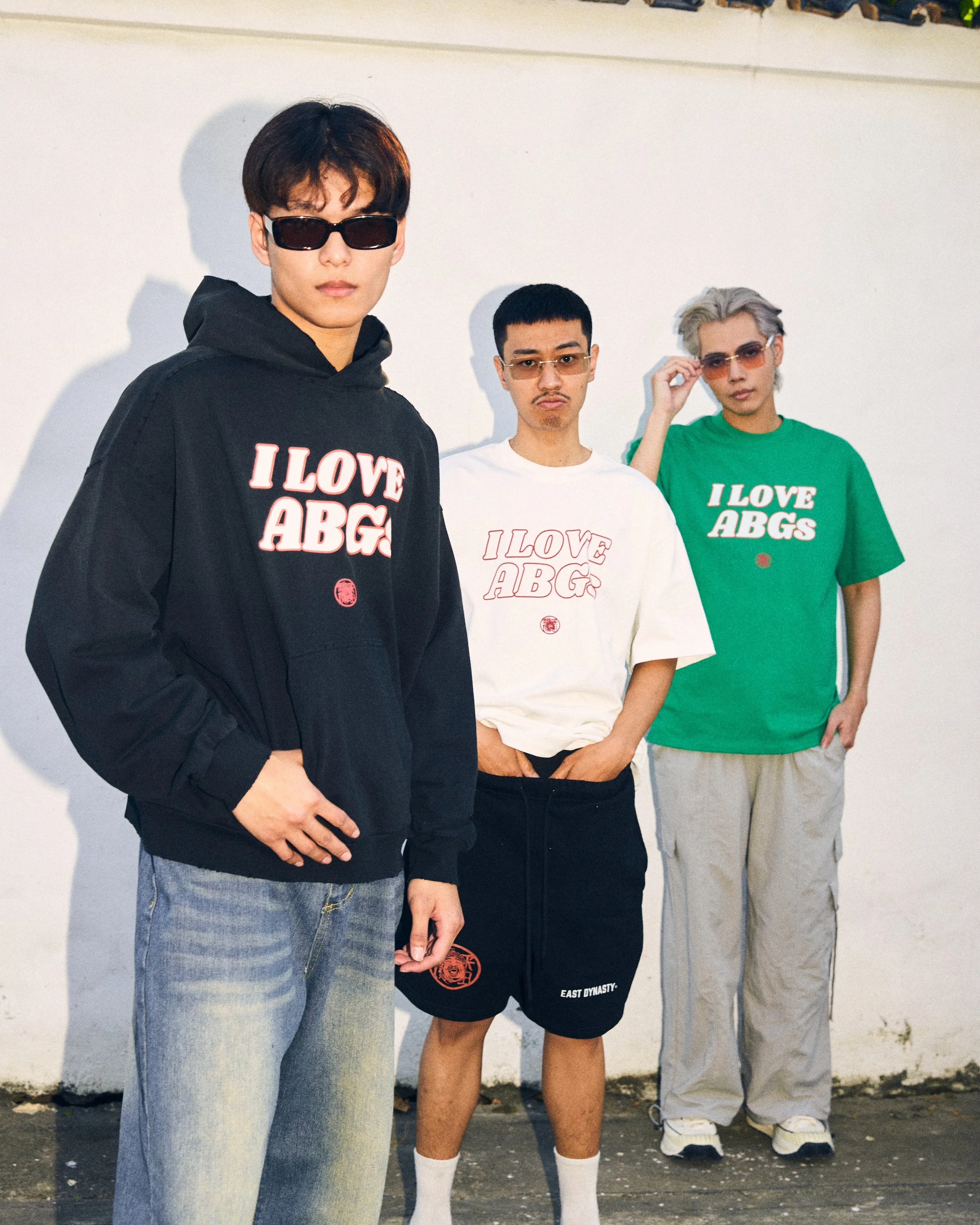 Three young men posing against a white wall wearing casual streetwear with sunglasses. The man on the left wears a black hoodie with red text that says 'I LOVE ABGS', ripped jeans. The man in the middle wears a white T-shirt with red outline text that says the same and black shorts. The man on the right wears a green T-shirt with white text that says the same, beige pants, and sunglasses.