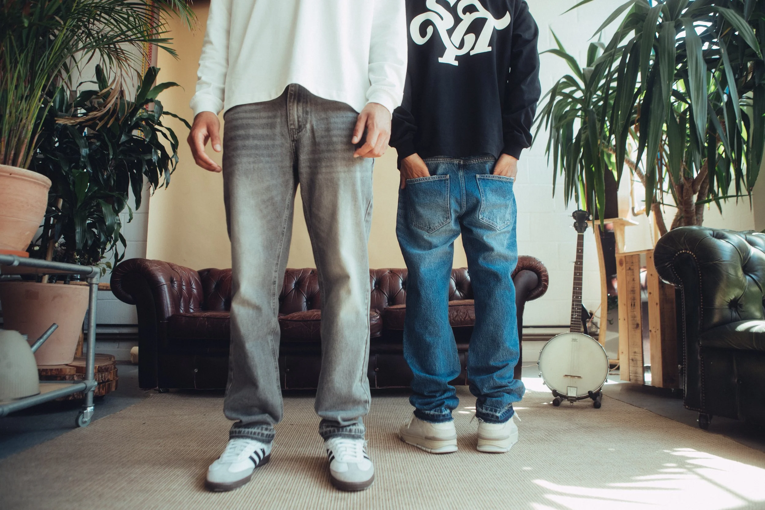 Two people standing indoors, only their lower bodies visible. The person on the left is wearing light gray jeans and white sneakers with black stripes. The person on the right is wearing blue jeans and white sneakers. The background includes a brown leather couch, large green plants, and a banjo on a stand.