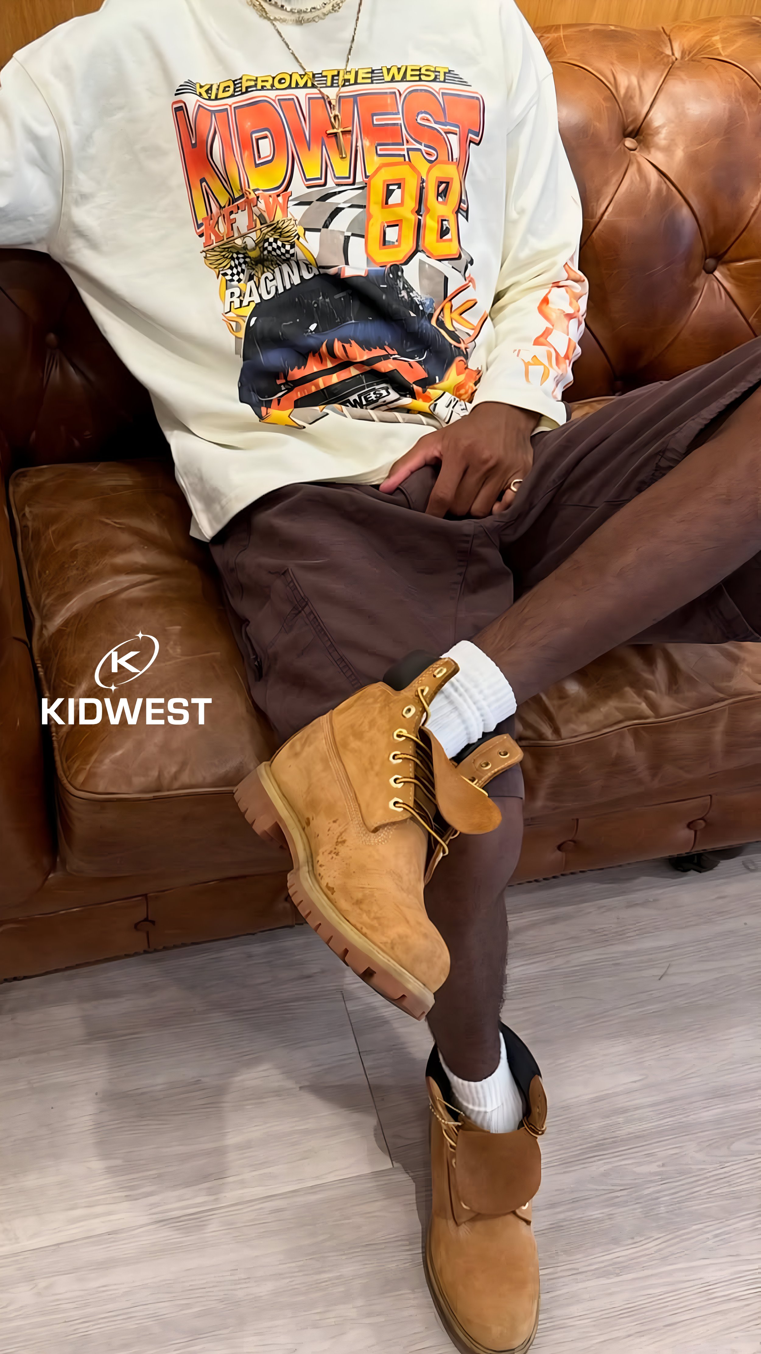 Person sitting on a brown leather couch wearing a colorful graphic T-shirt with racing imagery, dark shorts, white socks, and large tan work boots. The person's face is not visible.