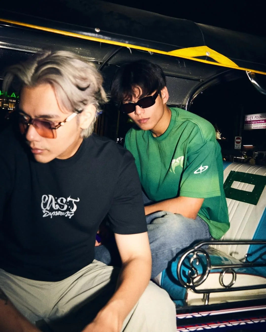 Two young men wearing sunglasses sitting in a tuk-tuk, one with blond hair and black shirt, the other with black hair and green shirt, at night.