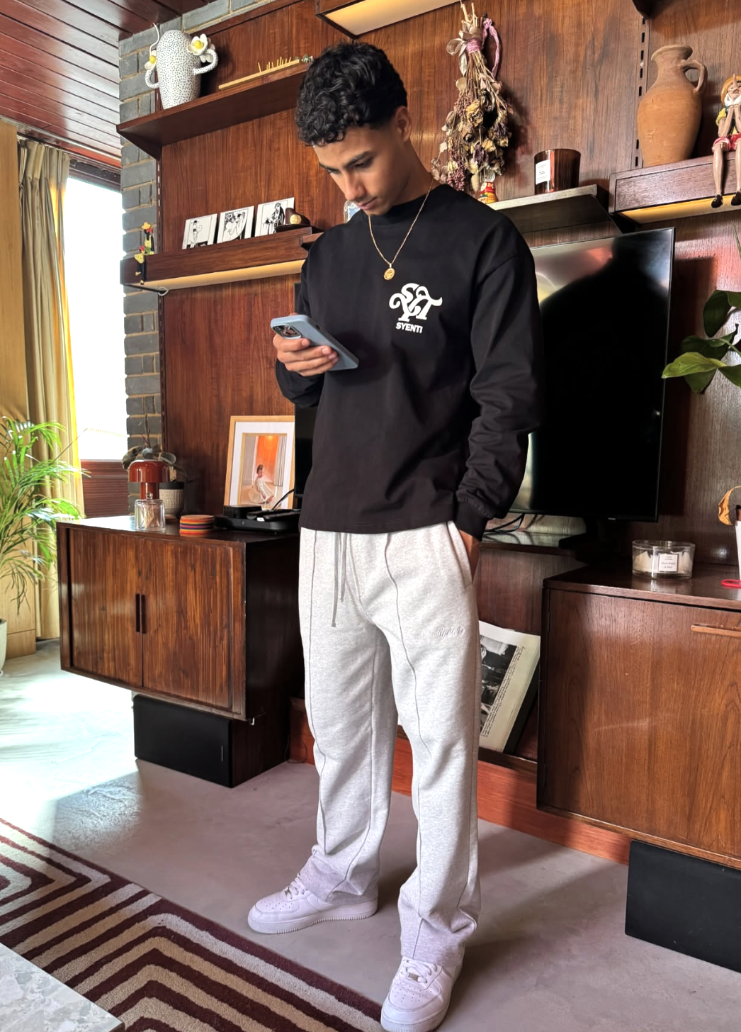 A young man with dark curly hair dressed in a black long-sleeve shirt and light gray sweatpants stands indoors looking at his phone. The background features a wooden wall with shelves, decorative art, potted plants, and a television.