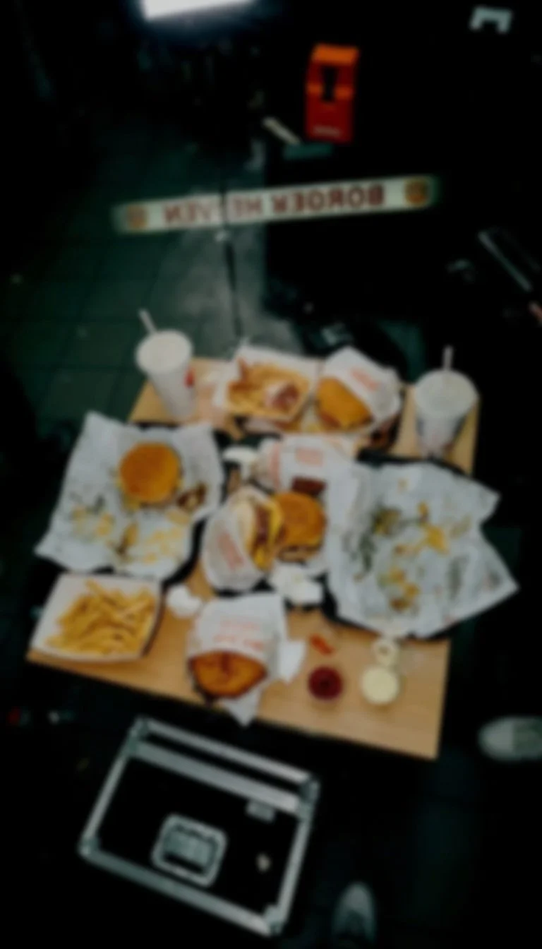 Fast food meal including onion rings, fries, sandwiches, drinks, and condiments on a small table across from a broken 'Burger King' sign.