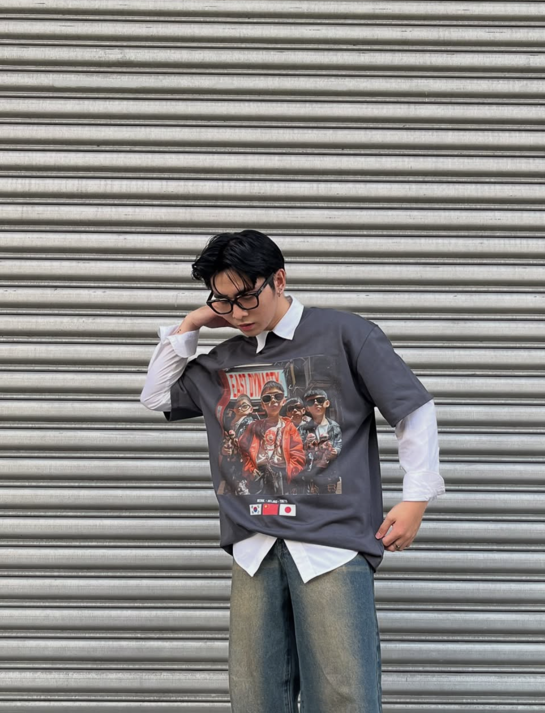 Young man with black hair, glasses, wearing a layered outfit with a streetwear graphic t-shirt over a white shirt, standing in front of metallic roller shutter.