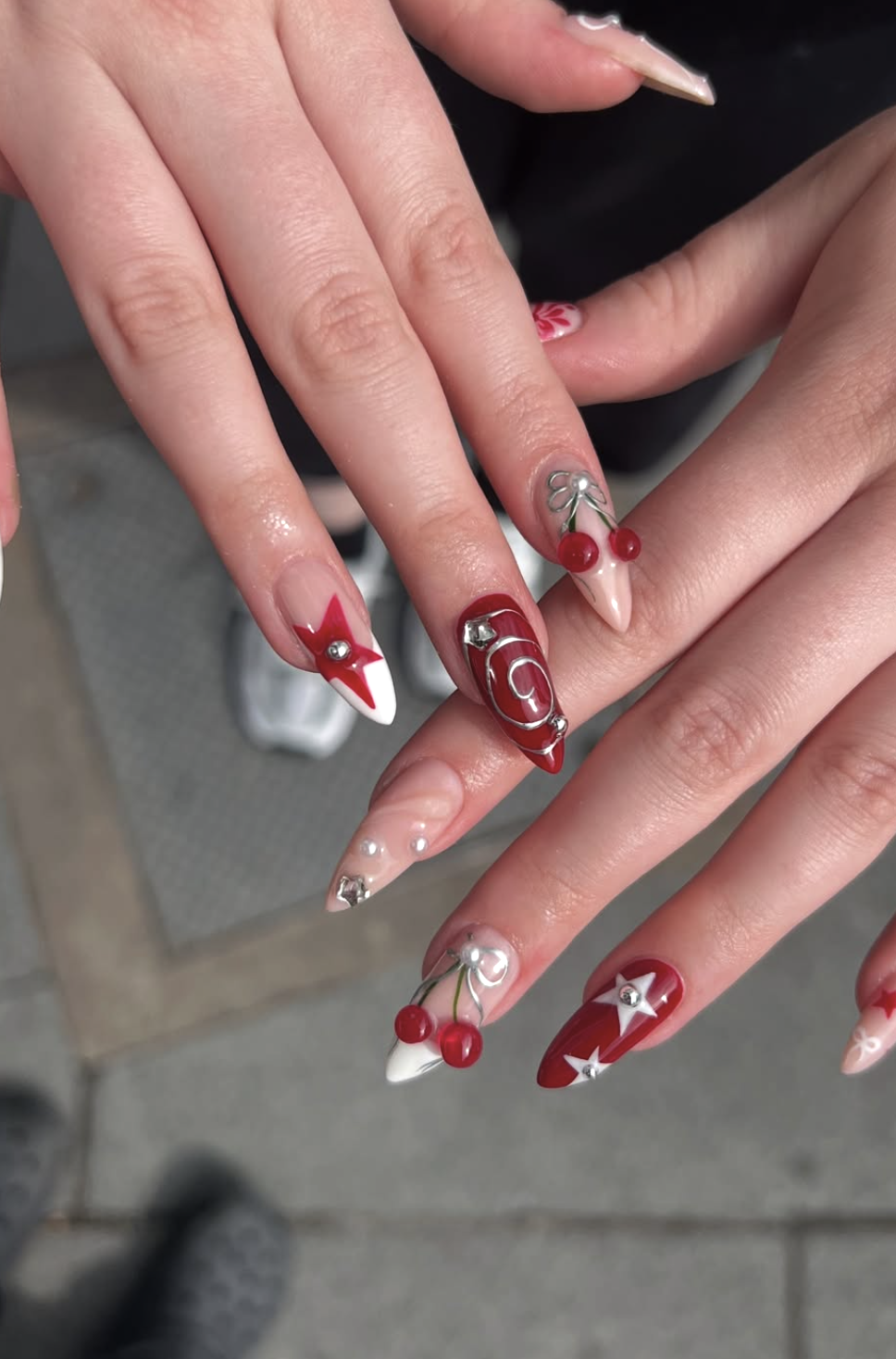 Close-up of hands with holiday-themed manicured nails featuring red, white, and silver colors, with decorations like cherries, stars, and swirling patterns.