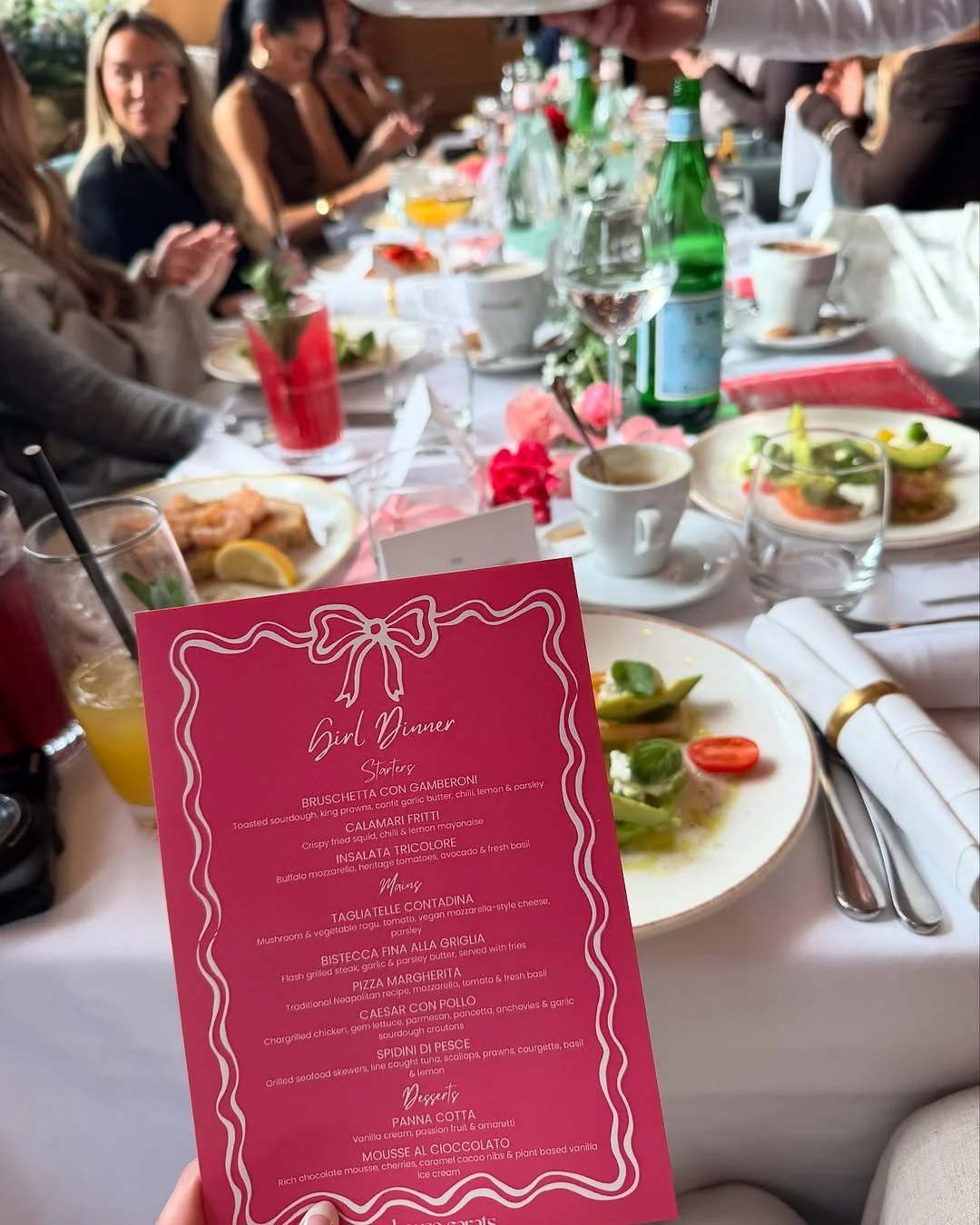 A table set for a girl dinner with various plates of food, drinks, and a pink menu card in the foreground.