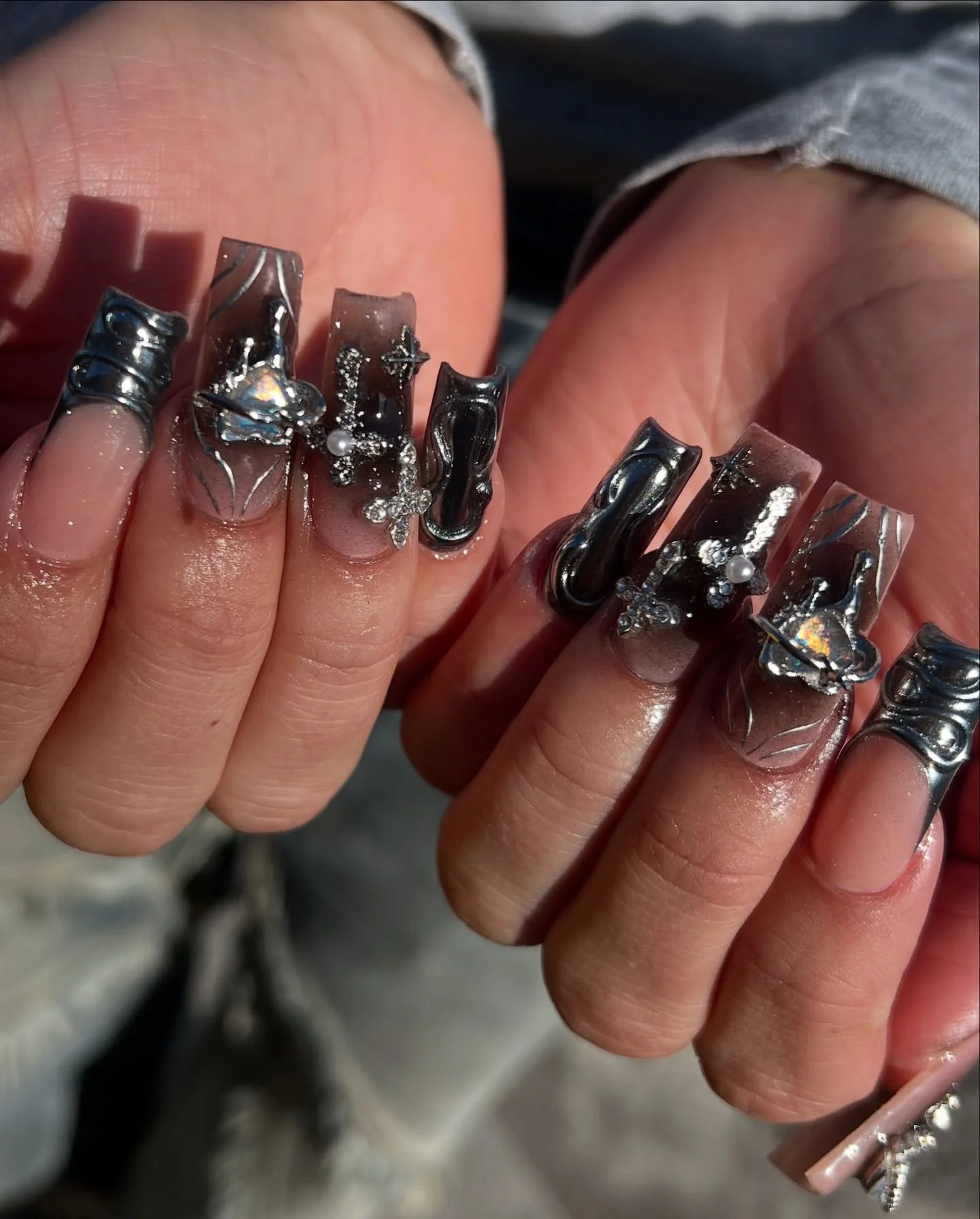 Close-up shot of a person's hands with elaborate acrylic nail art, featuring metallic, swirl, and decorative embellishments including small pearls and rhinestones.