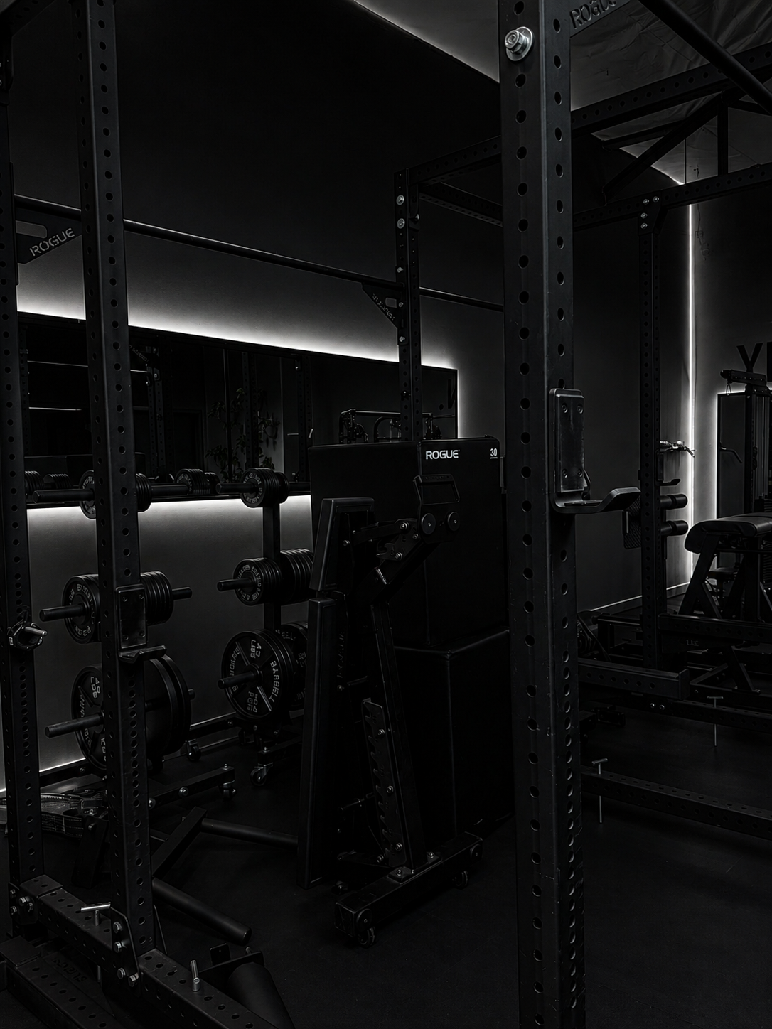 A gym with black weightlifting equipment, including barbells and weight plates, in a dark room with minimal lighting.