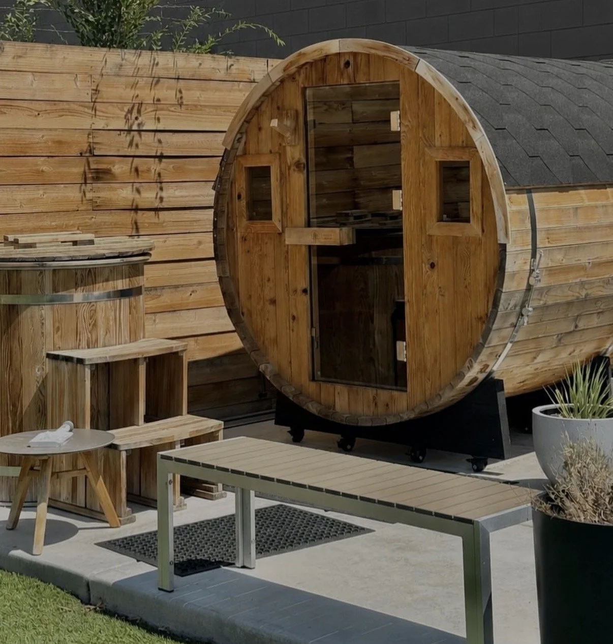 A wooden barrel sauna with a glass door and small windows, situated in an outdoor space with wooden fencing, potted plants, a small table, a bench, and a lounge chair on a concrete patio.