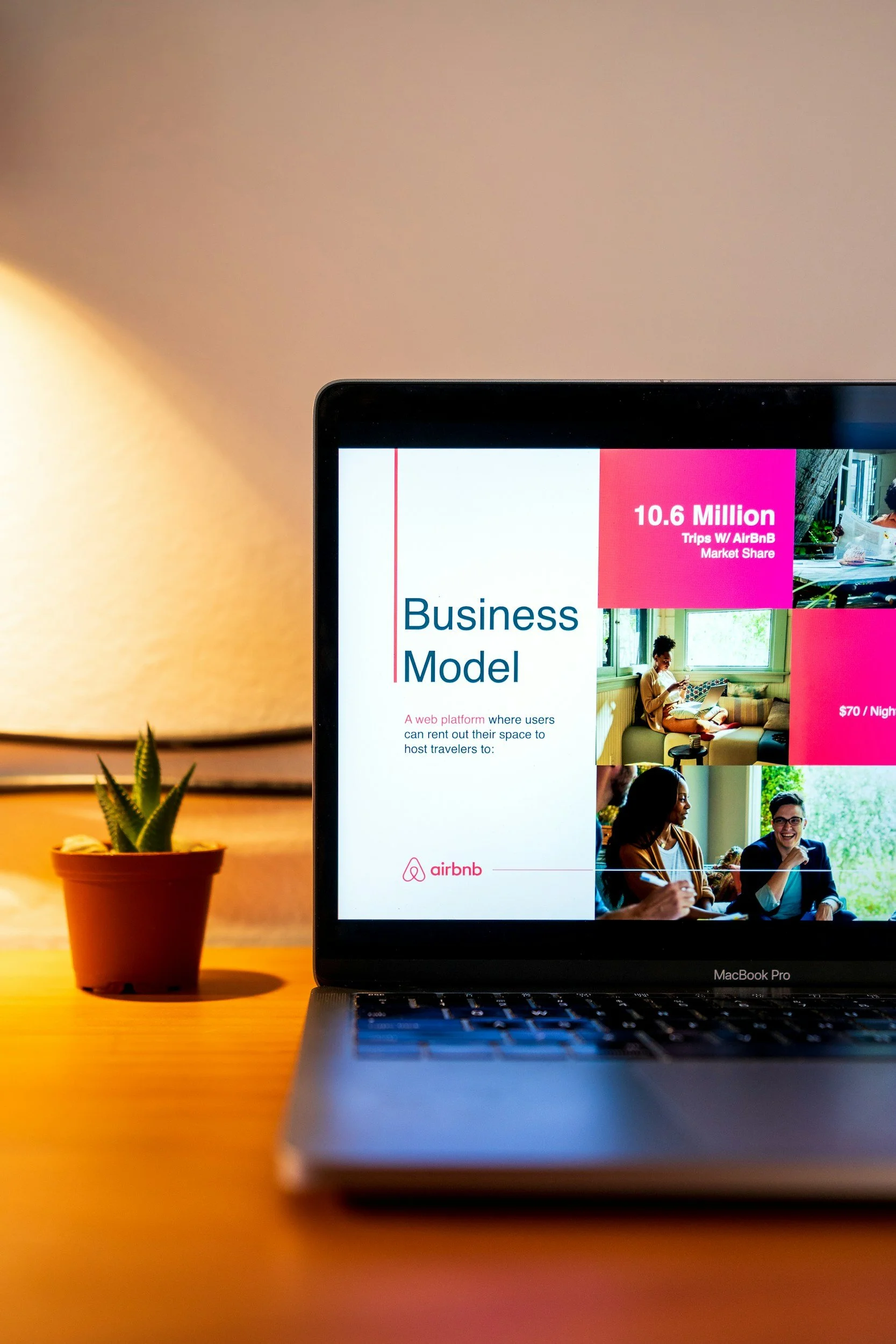 Part of a laptop screen displaying Airbnb business model with a pink and white presentation slide, a woman using a laptop, and people in a cozy sunny room. A small potted succulent plant is on the wooden desk.