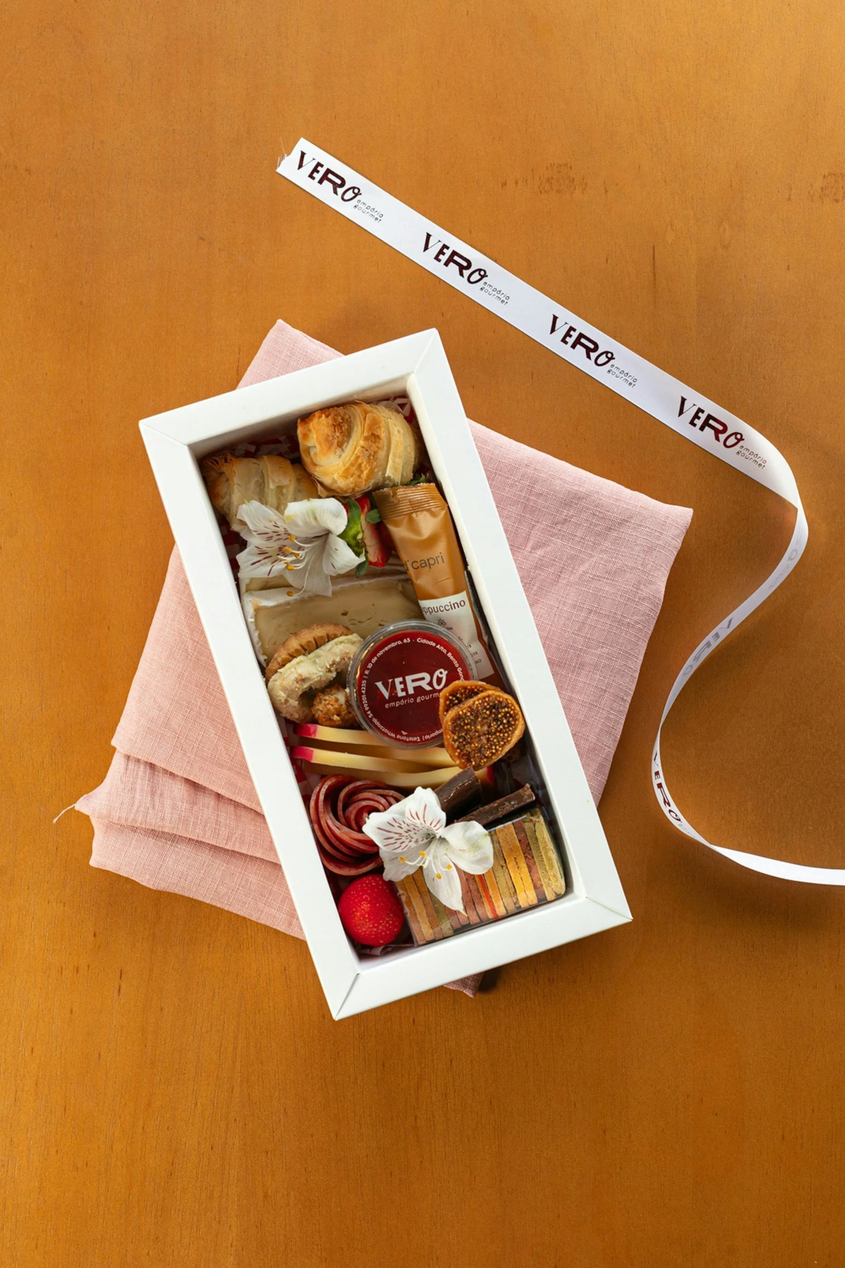 A white rectangular box filled with an assortment of desserts and chocolates, decorated with white flowers, placed on a pink cloth on a wooden surface.