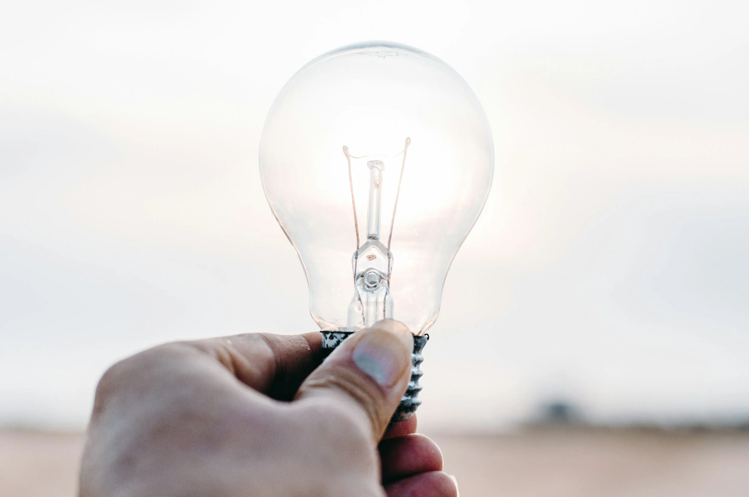 Close-up of a hand holding a clear incandescent light bulb against a light sky background.
