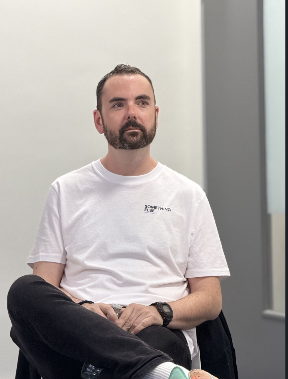 A bearded man sitting with his legs crossed in a white t-shirt that says 'SOMETHING ELSE', wearing a watch, in front of a plain wall.