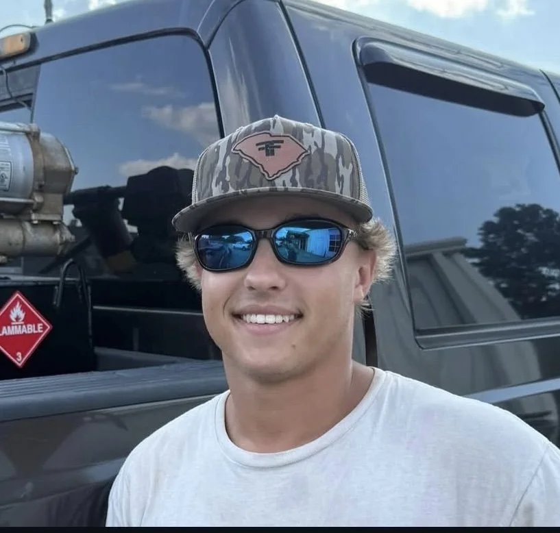 Young man smiling outdoors, wearing a camouflage baseball cap with a state outline and logo, a camouflage and brown jacket, standing in front of a pickup truck on a sunny day with blue sky and trees in the background.