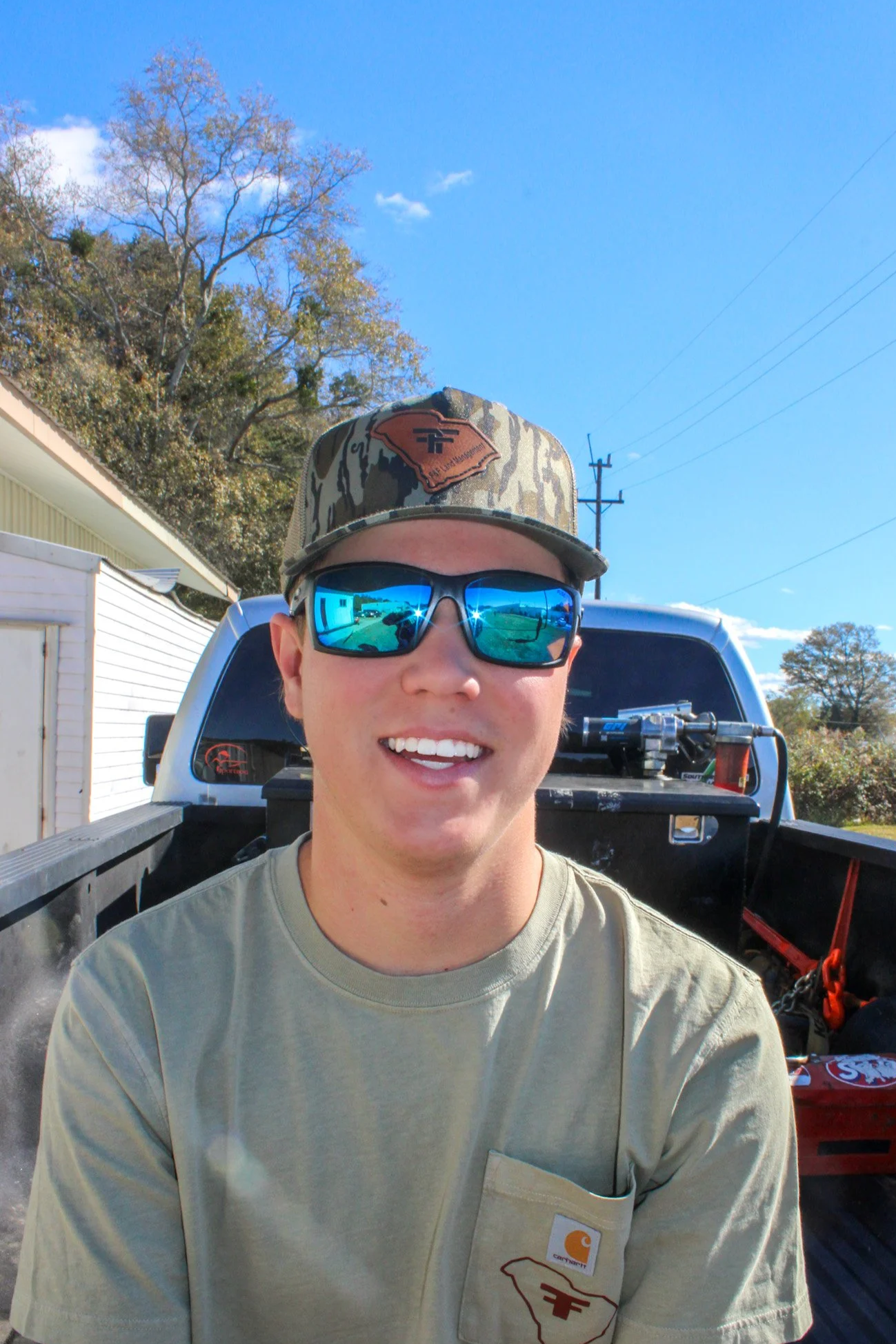 A young man smiling and wearing sunglasses, a camouflage cap with a patch, and a beige t-shirt, sitting in the back of a pickup truck on a sunny day with blue sky, trees, utility poles, and a building in the background.