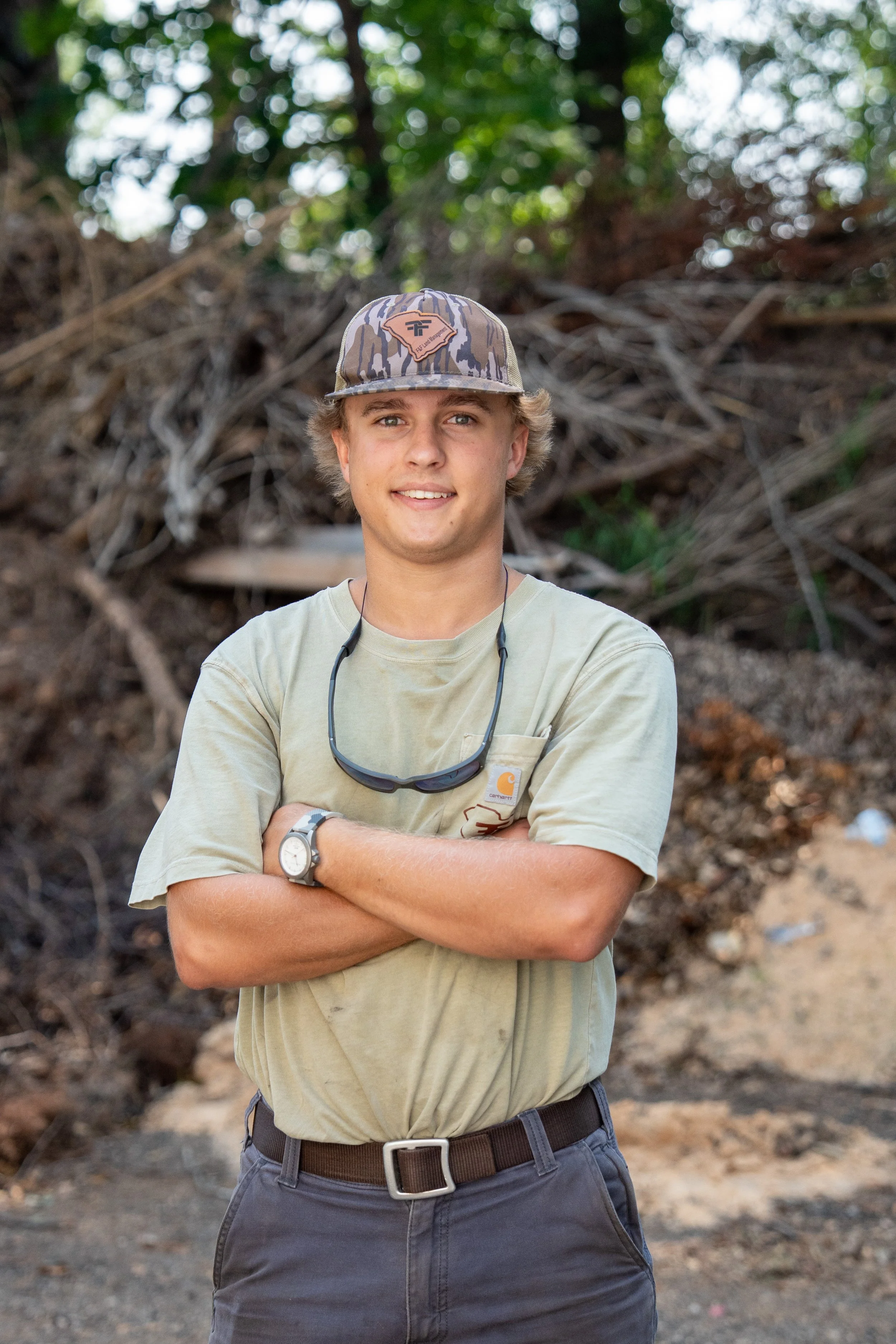 Young man smiling outdoors, wearing a camouflage baseball cap with a state outline and logo, a camouflage and brown jacket, standing in front of a pickup truck on a sunny day with blue sky and trees in the background.