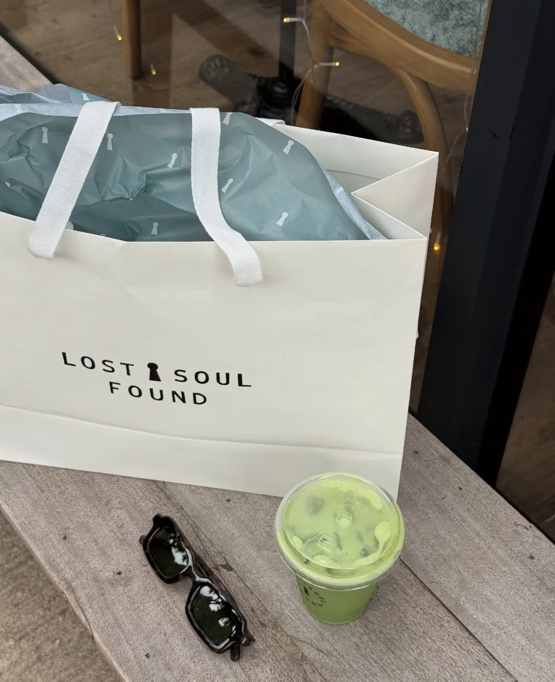 A white paper bag with the words 'Lost Soul Found' printed on it, a pair of black sunglasses, and a green iced drink on a wooden surface.