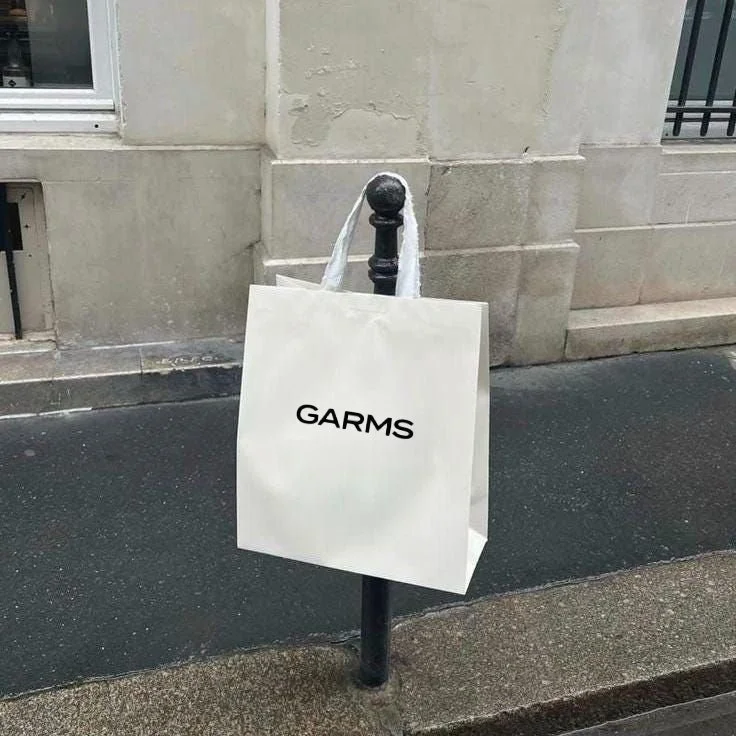 A white shopping bag hanging on a black street post with the text "GARMS" written on it, located on a city sidewalk with a building wall in the background.