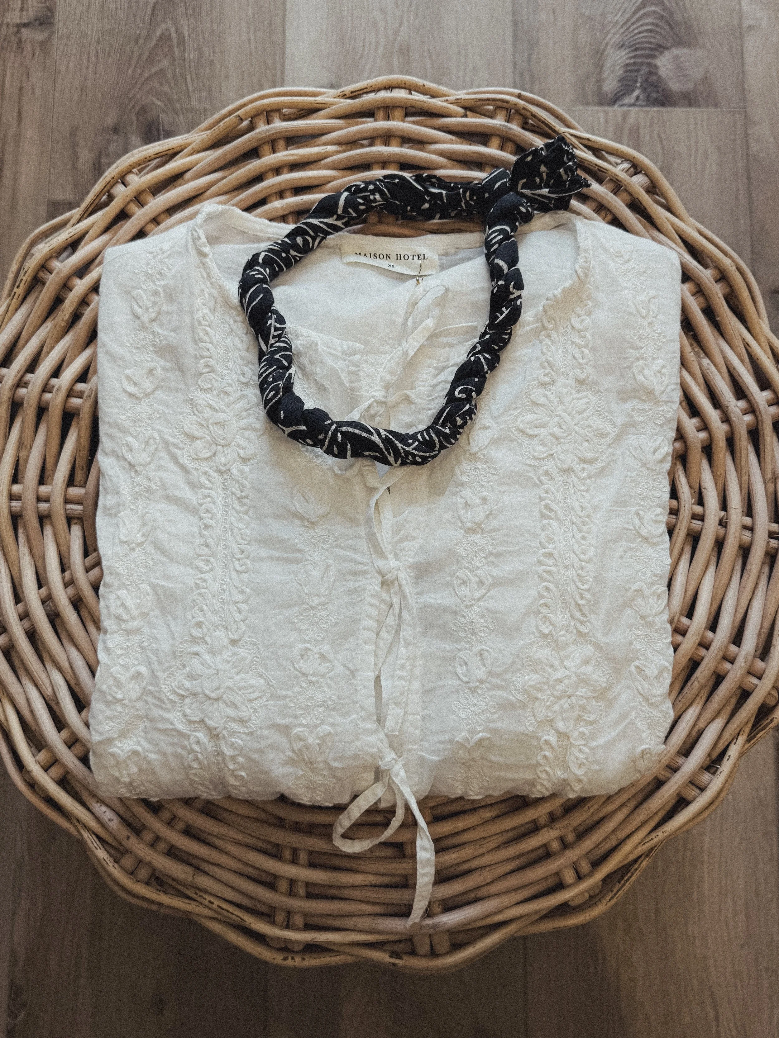 A folded white embroidered blouse with a tassel tie in the front, placed on a wicker basket, with a black and white patterned scarf around the collar.