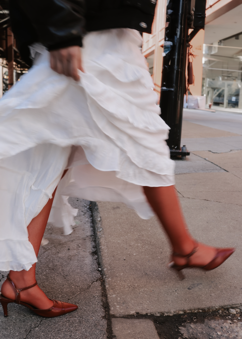 A person walking on a city sidewalk wearing a white ruffled dress, reddish-brown stockings, and matching reddish-brown high heels, holding the dress with one hand, with storefronts and sidewalk details visible.