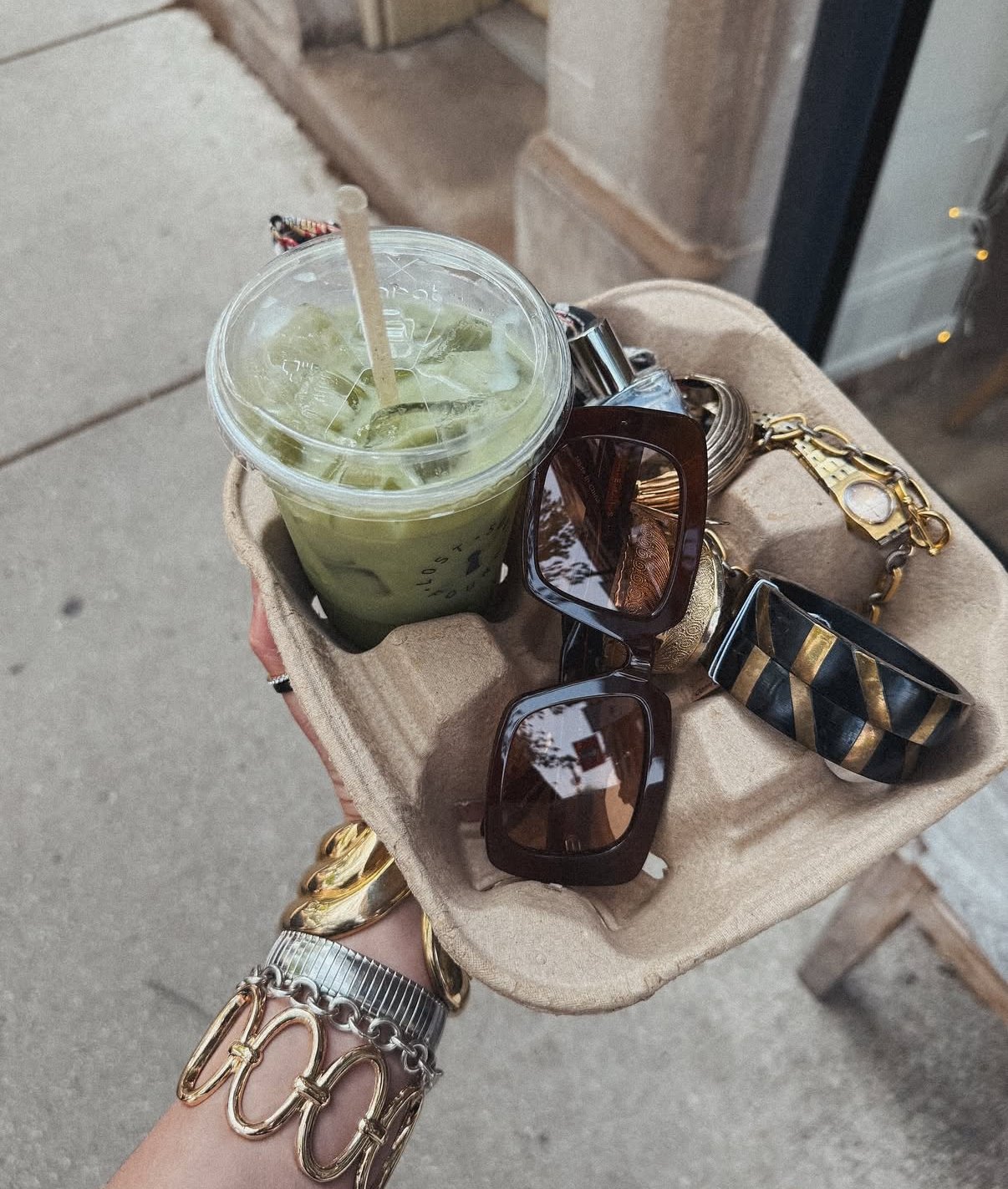 A hand holding a tray with a green iced drink and several accessories including sunglasses, bracelets, and jewelry, outdoors on a sidewalk.