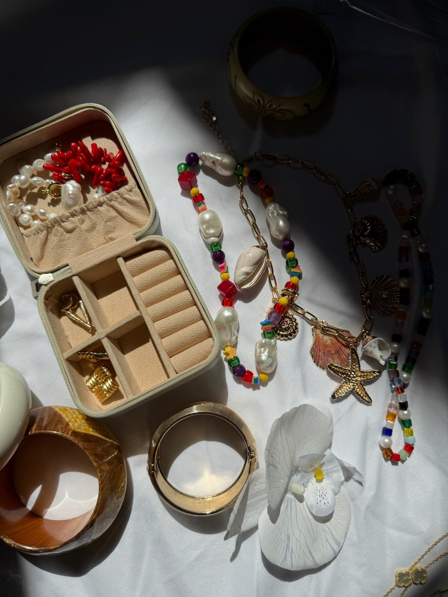 Assorted jewelry including necklaces, bracelets, rings, and earrings, some with seashell and starfish charms, arranged on a white surface with a jewelry box and a white orchid flower.