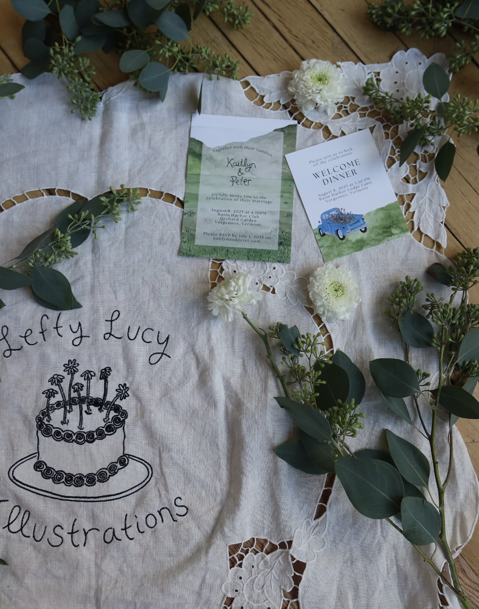 Wedding invitation and welcome dinner card surrounded by white flowers, green foliage, and a embroidered cloth with a birthday cake illustration.