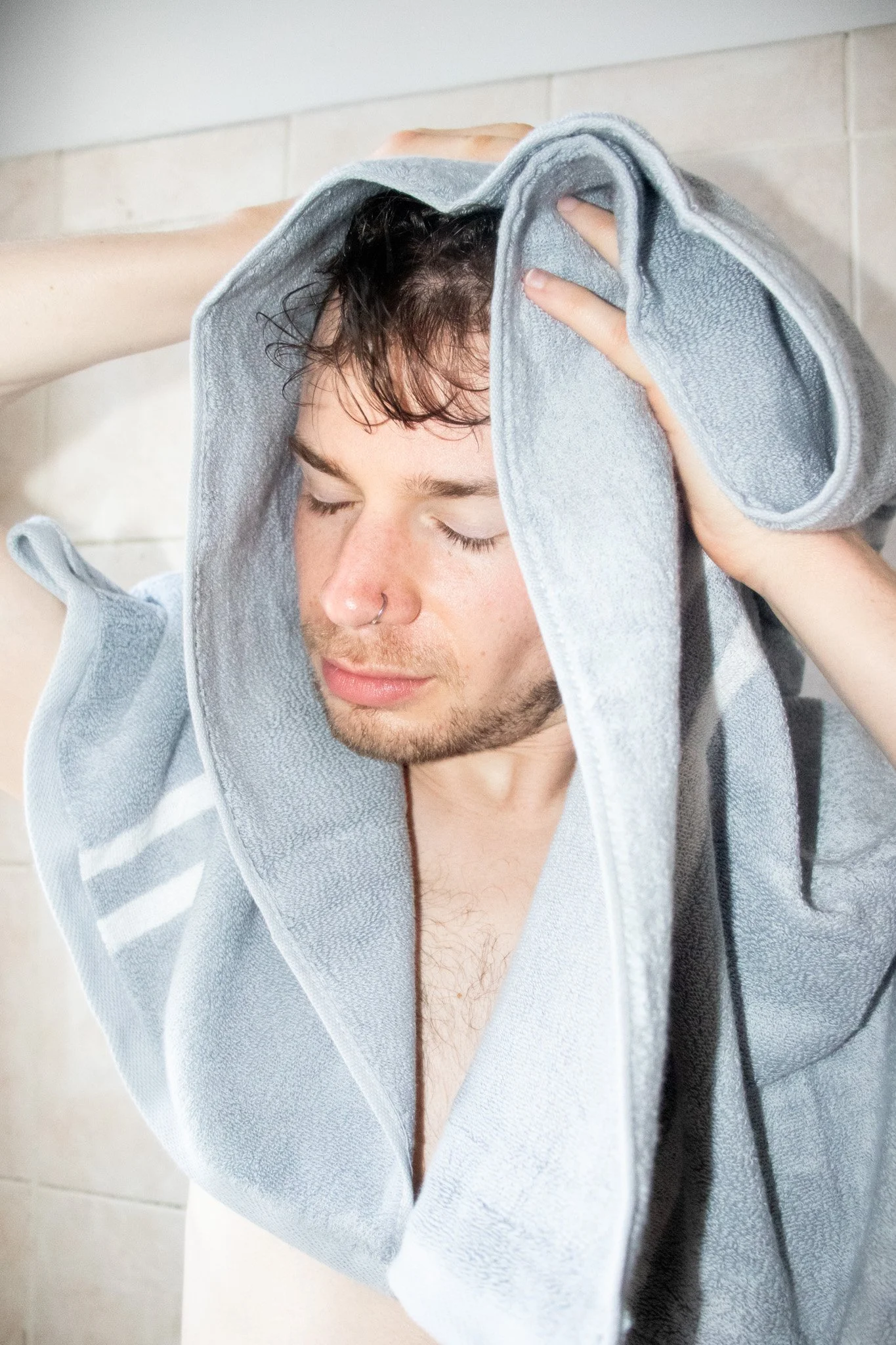 A young man with wet hair and a nose piercing drying his head with a gray towel after a shower.