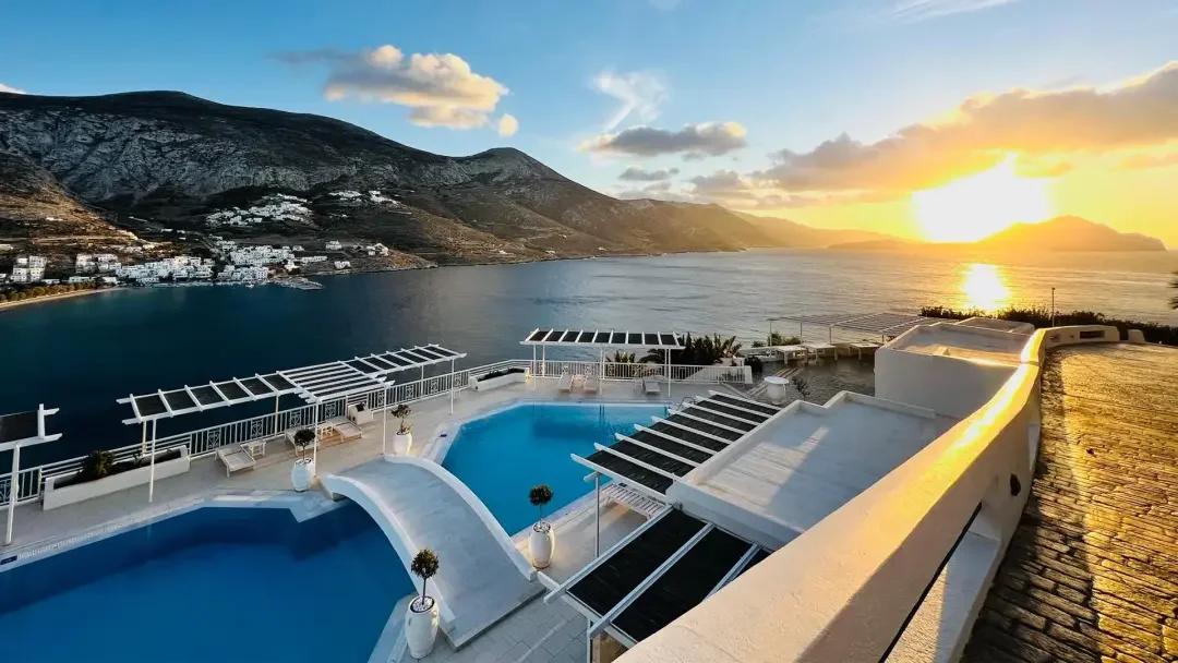 Sunset over a terrace with two small swimming pools, overlooking a bay with mountains in the background, and a partly cloudy sky.