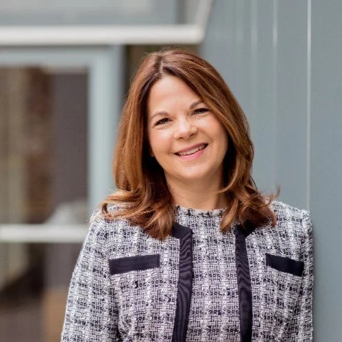 Woman with shoulder-length brown hair smiling, wearing a black and white patterned blazer, standing outside near a glass building.