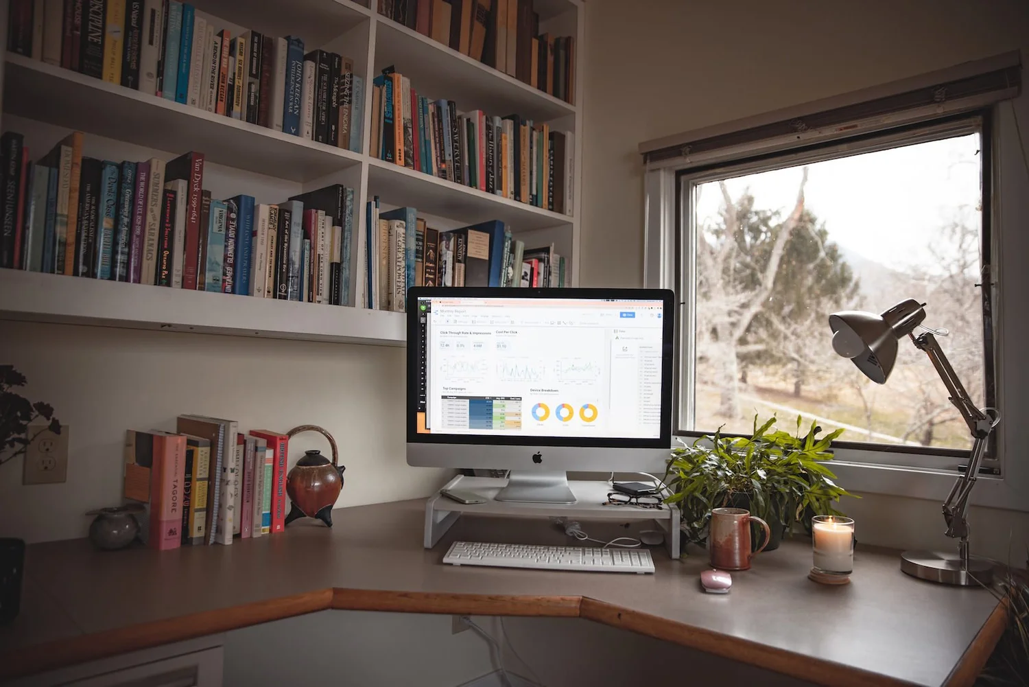 A home office desk with a computer showing a marketing analytics dashboard, beside a window and bookshelves