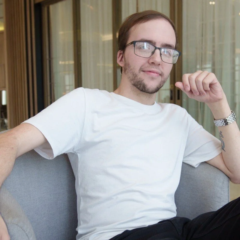 A young man sitting on a gray couch indoors, wearing glasses, a white t-shirt, and a watch, with a modern glass-walled room in the background.