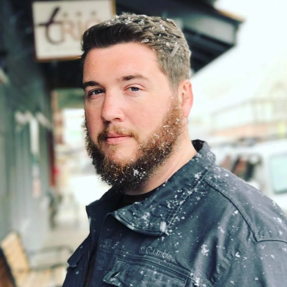 A man with a beard and short brown hair standing outdoors on a snowy day, wearing a black Columbia jacket with snow on his shoulders and face, looking at the camera with a slight smile.