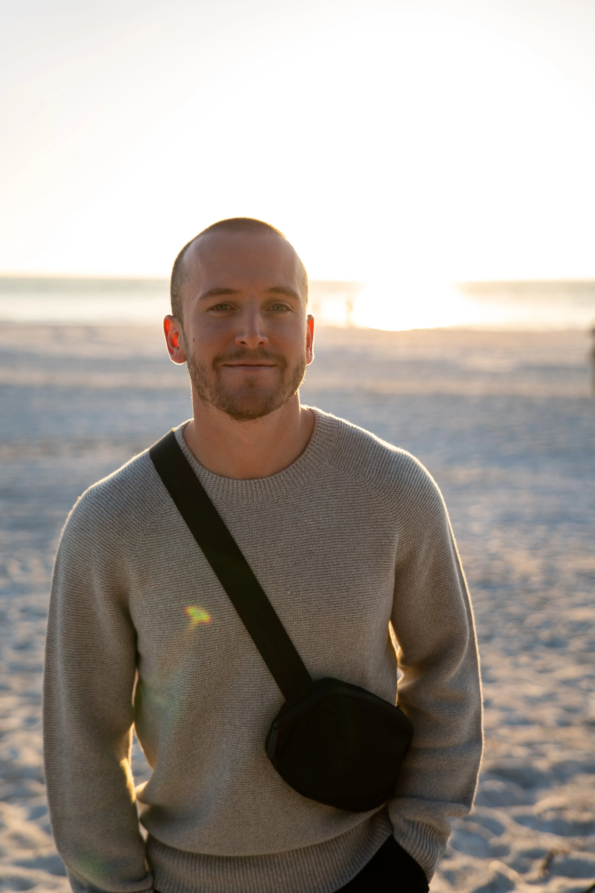 Junger Mann mit kurzhaarigem Bart und einem hellgrauen Pullover, trägt einen schwarzen Schultergurt, steht am Strand bei Sonnenuntergang, im Hintergrund das Meer und die Sonne.