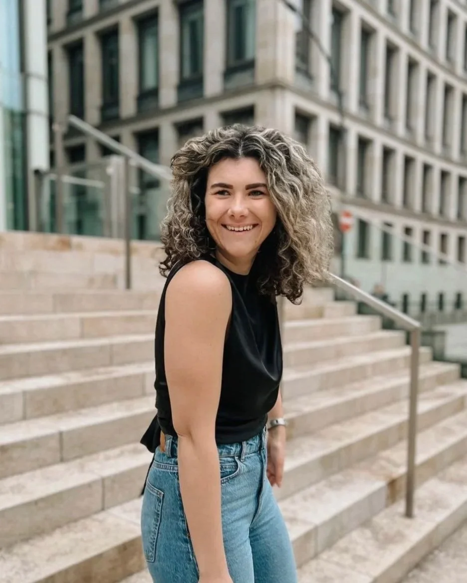 Junge Frau mit lockigem Haar, schwarze Top, blaue Jeans, lächelnd, auf Treppen vor einem modernen Gebäude in der Stadt.