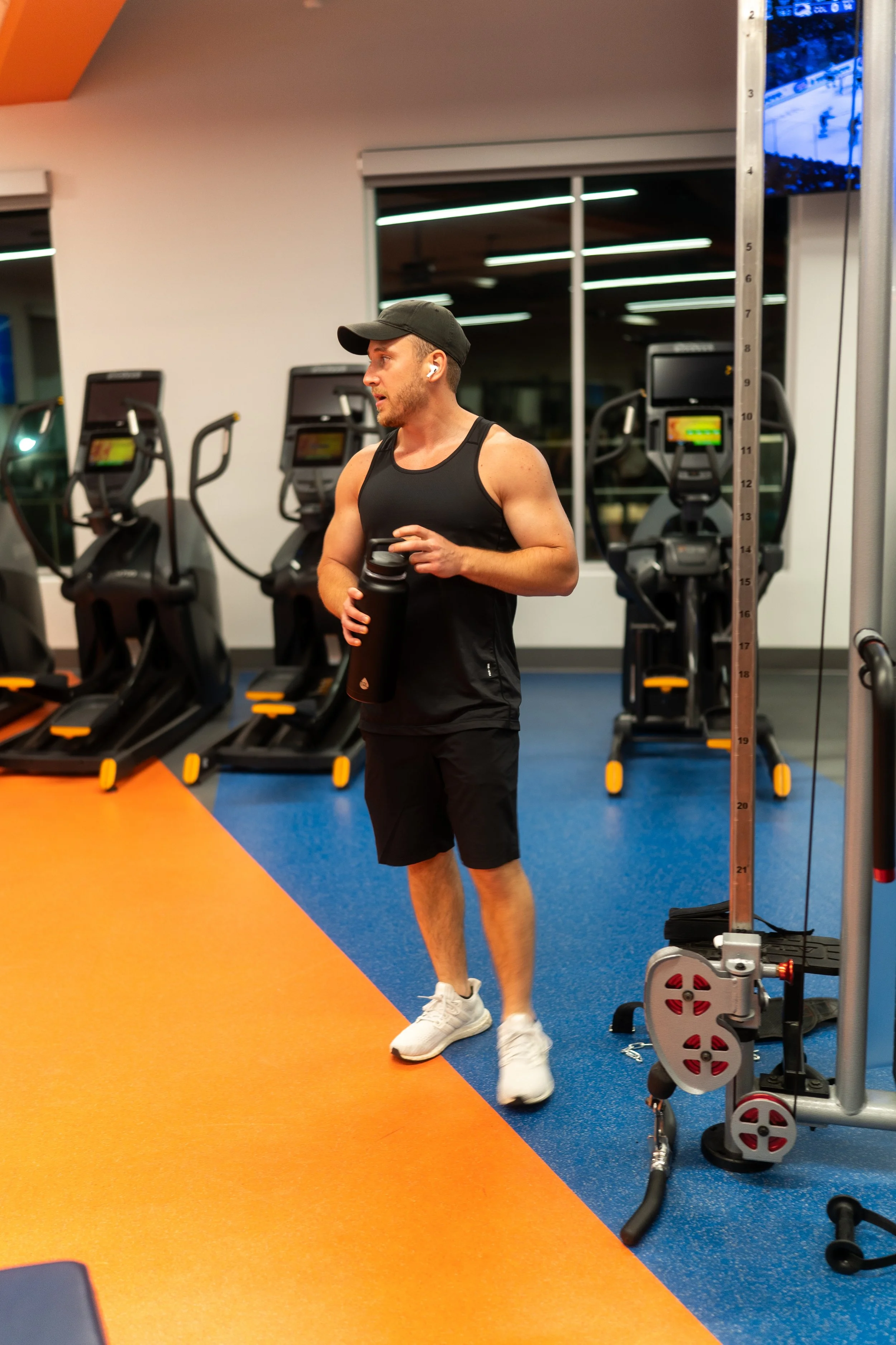 Mann beim Training im Fitnessstudio, trägt schwarze Sportkleidung, eine schwarze Kappe und benutzt eine Wasserflasche. Im Hintergrund sind Laufbänder und ein großes Fenster, das in einen Raum mit Deckenlichtern blickt.