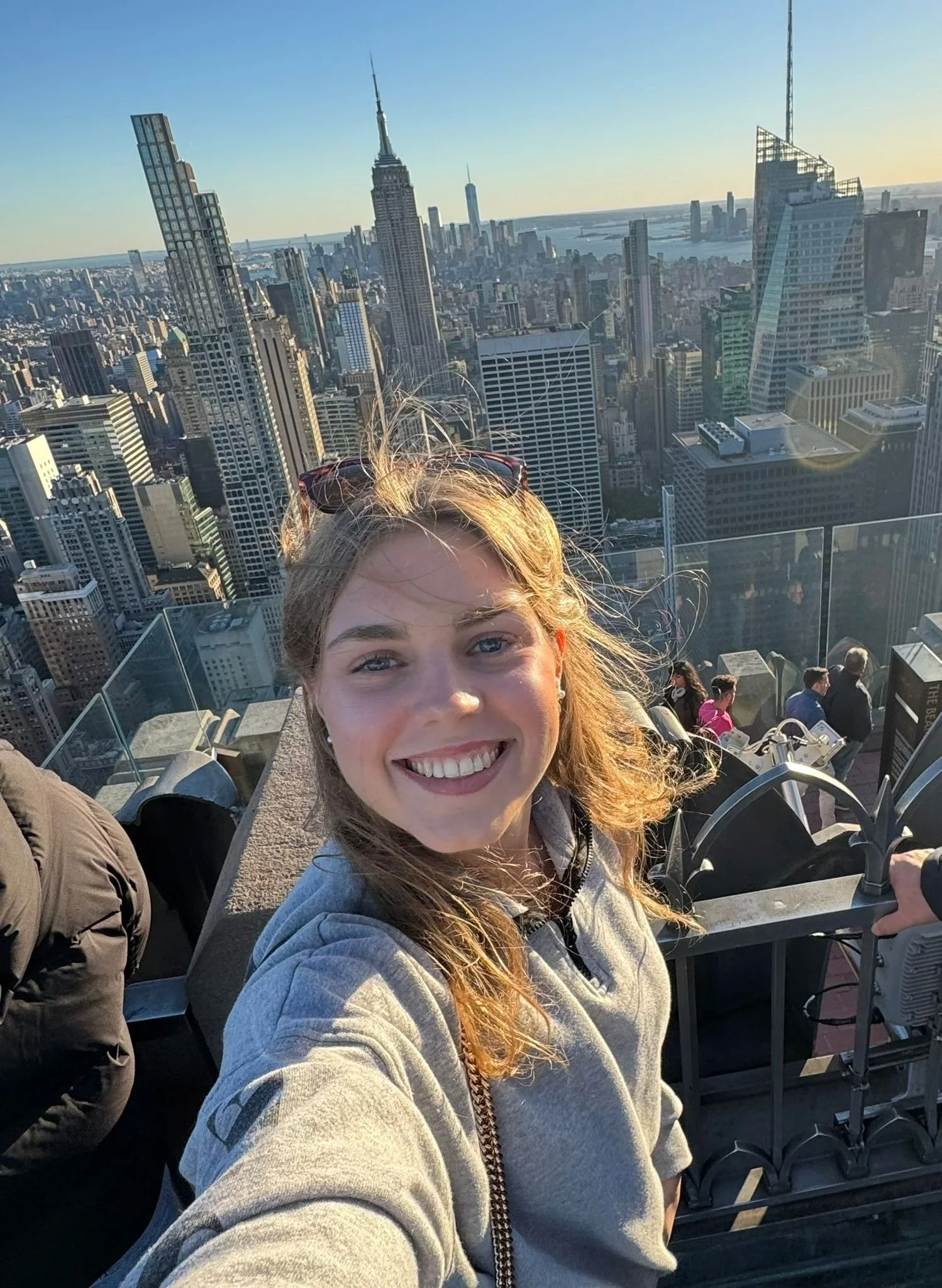 Freudige junge Frau mit langen, blondem Haar macht Selfie auf Aussichtsplattform in New York City mit Blick auf die Skyline, inklusive Empire State Building, bei Sonnenuntergang, im Hintergrund sind andere Touristen zu sehen.