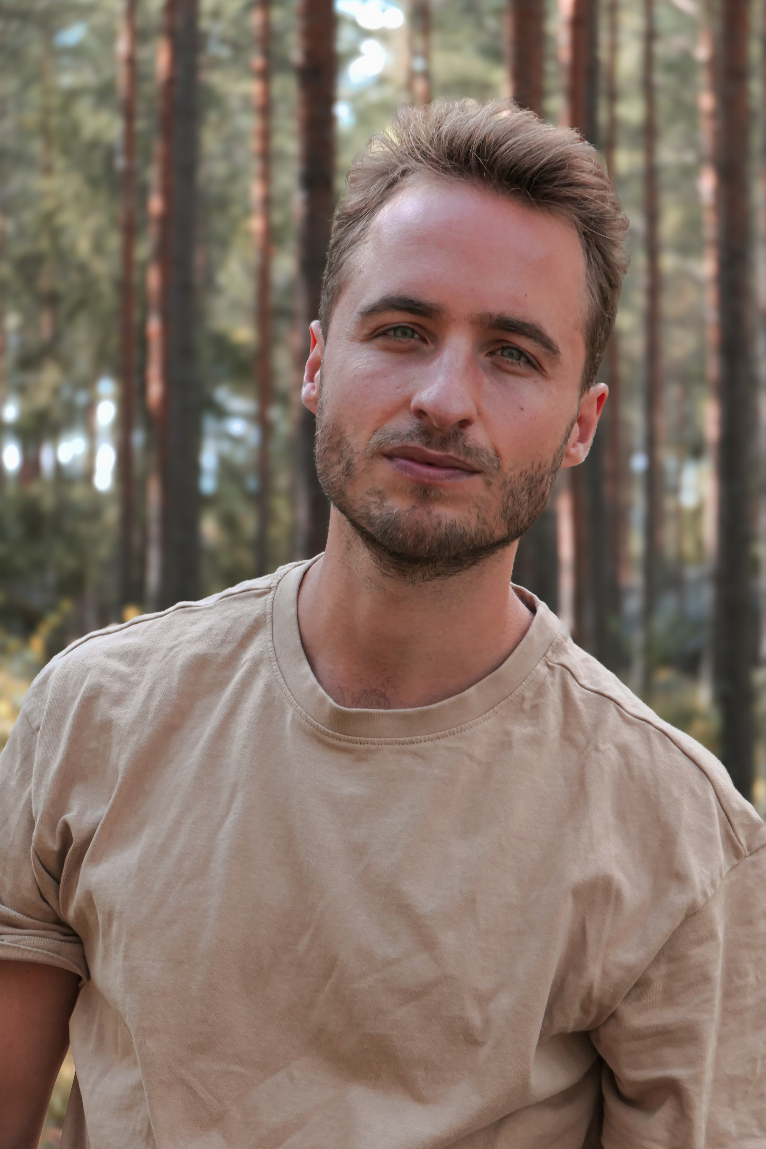 Ein junger Mann mit braunen Haaren und Bart trägt ein beige T-Shirt und steht im Wald zwischen Bäumen.