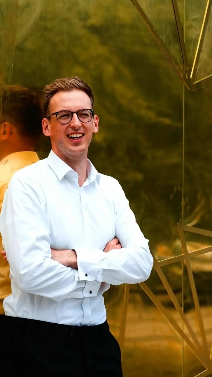 Ein junger Mann mit Brille und weißem Hemd, lächelnd, steht vor einer goldenen Wand im Hintergrund.