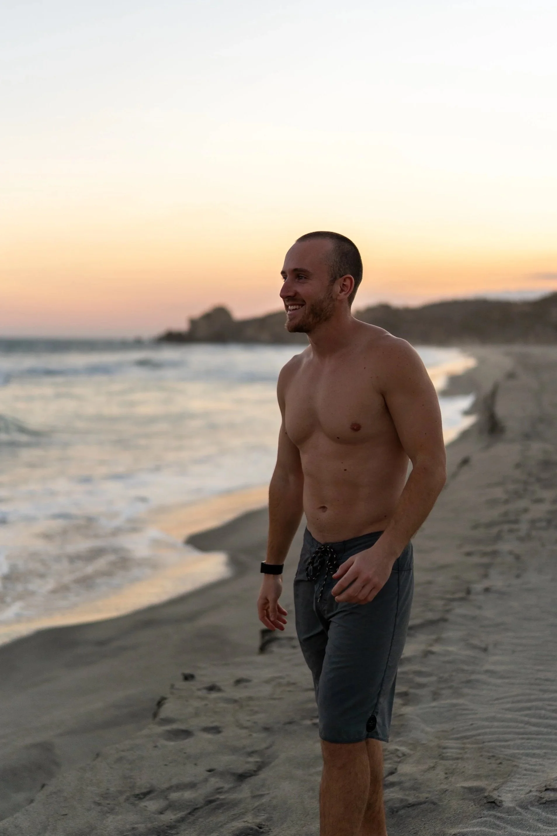 Ein lächelnder junger Mann ohne Hemd, trägt Shorts und steht am Strand bei Sonnenuntergang, mit Sand und Wasser im Hintergrund.