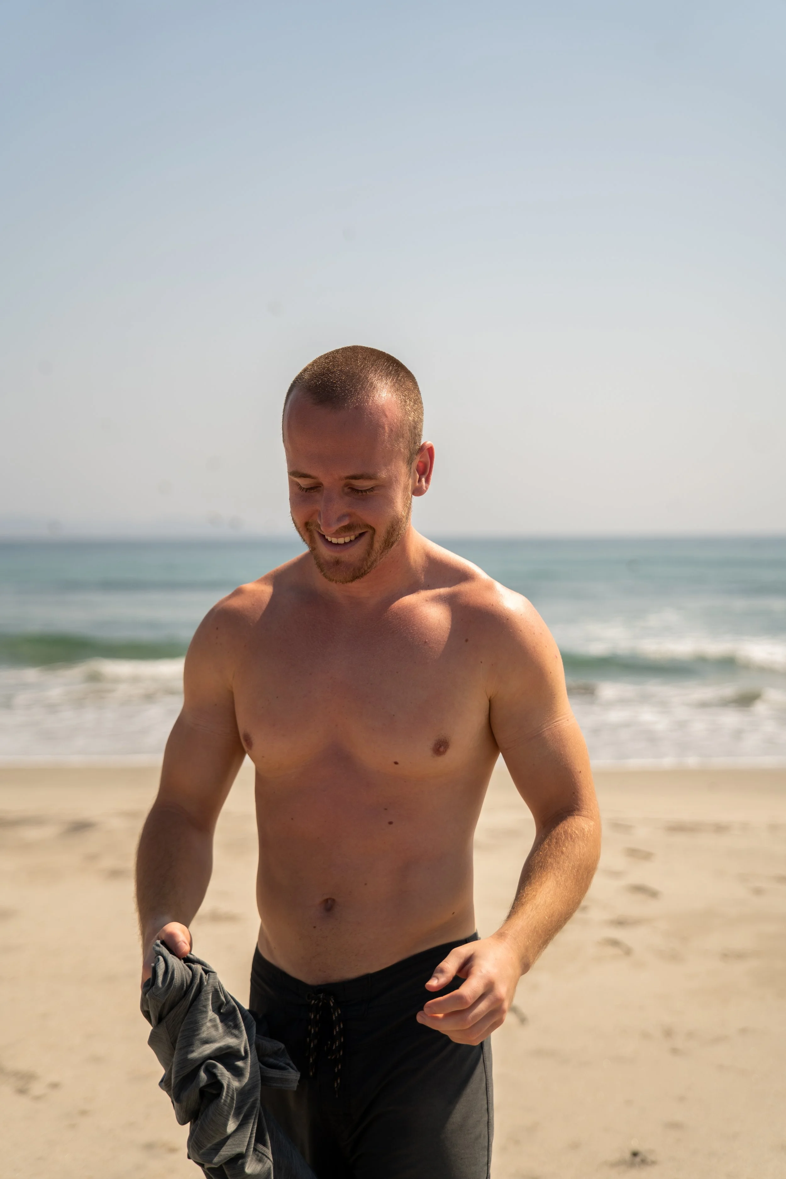 Ein lächelnder, muskulöser Mann ohne Oberkörper auf einem Strand, hält ein Kleidungsstück in der Hand, mit Meer im Hintergrund, bei sonnigem Wetter.