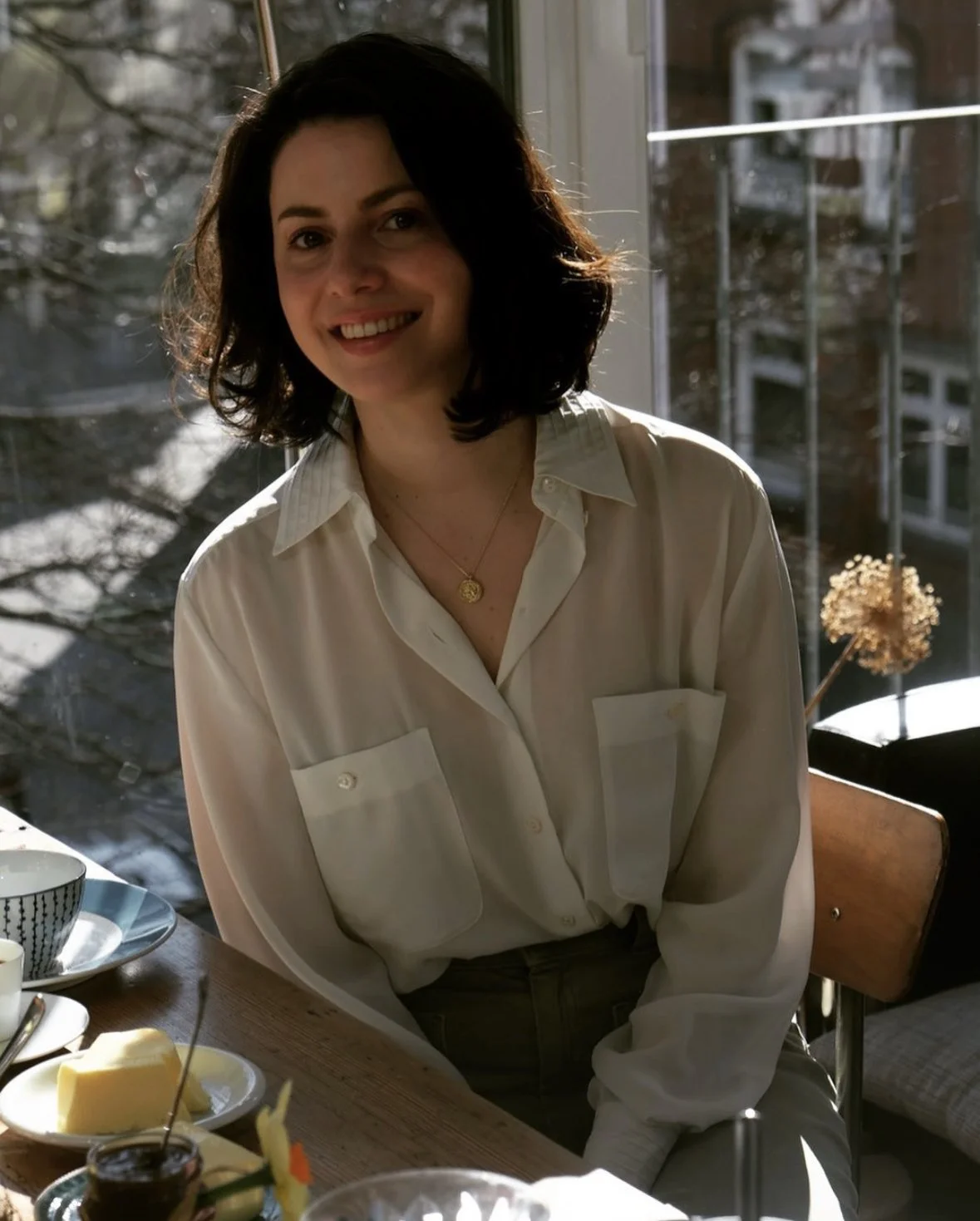 Eine lächelnde Frau mit schwarzen, lockigen Haaren, trägt ein weißes Hemd und sitzt an einem Tisch mit Frühstück am Fenster, im Hintergrund sind Bäume sichtbar.