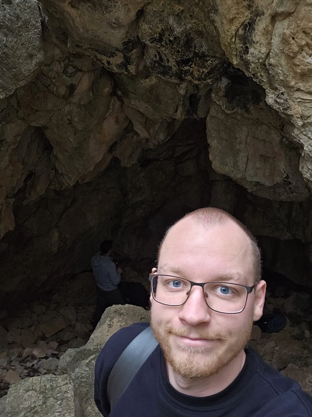 Ein Mann mit Brille und Bart macht ein Selfie vor einer Felsspalte in einer Höhle mit zwei weiteren Personen im Hintergrund.