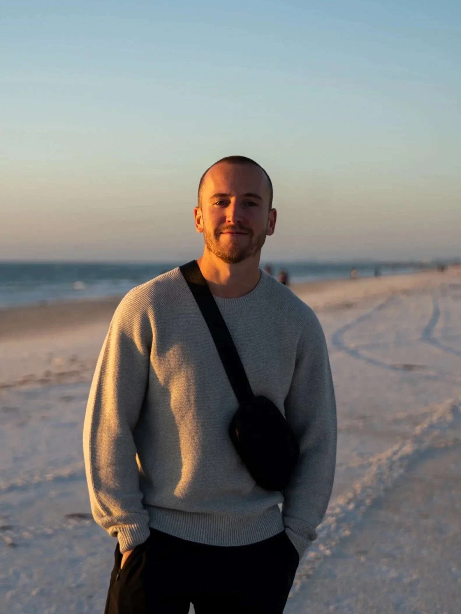 Ein Mann steht am Strand bei Sonnenuntergang, trägt ein graues Sweatshirt und eine schwarze Tasche, im Hintergrund Meer und Himmel.