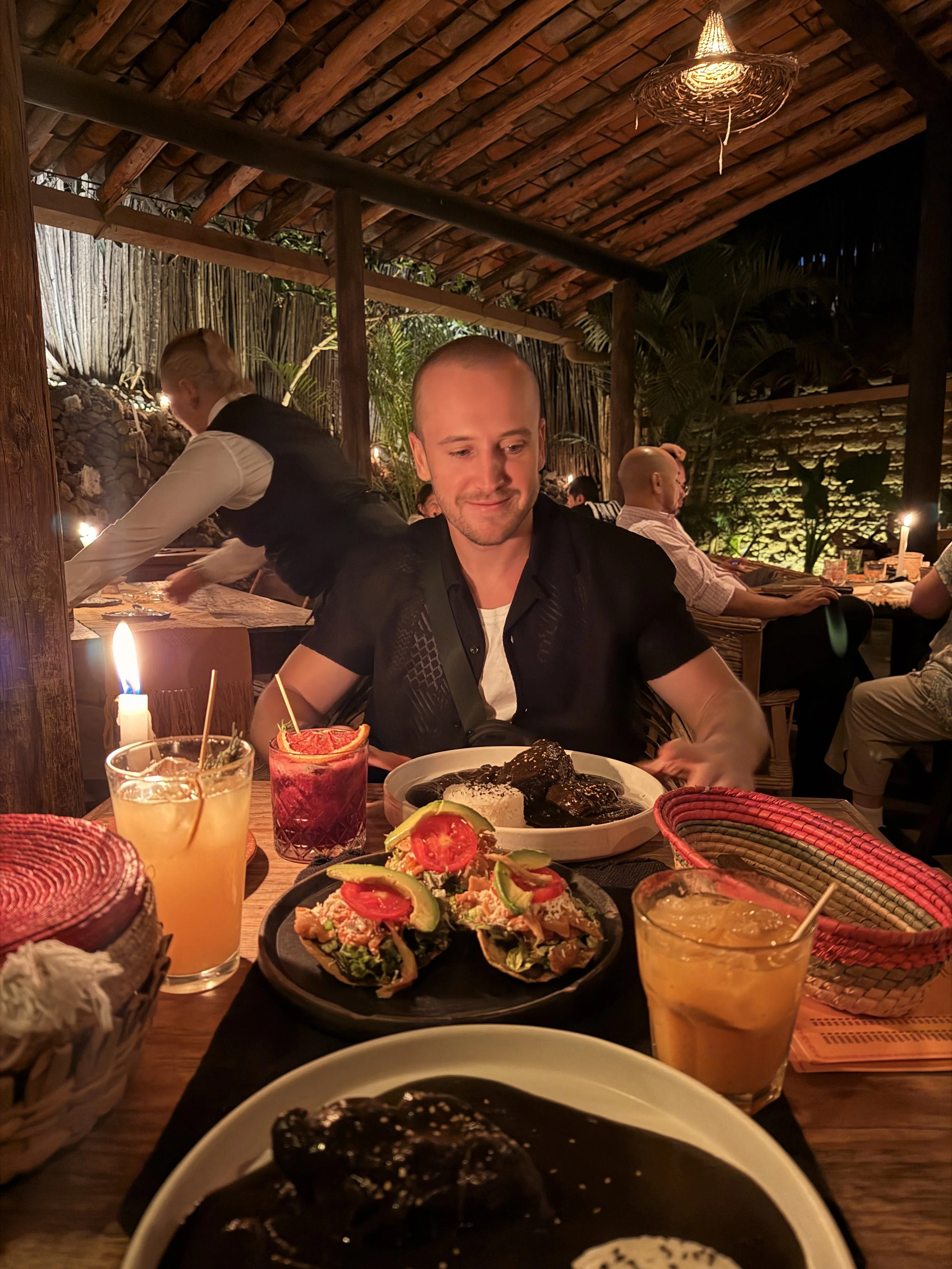 Ein Mann, der in einem Restaurant sitzt, es gibt mexikanisches Essen auf dem Tisch, darunter Enchiladas mit Avocado und Tomaten. Es sind Getränke vor ihm, darunter ein orangefarbener Drink und ein roter Drink mit Zitrone. Im Hintergrund sind weitere Gäste und eine dekorative, holzverkleidete Decke.