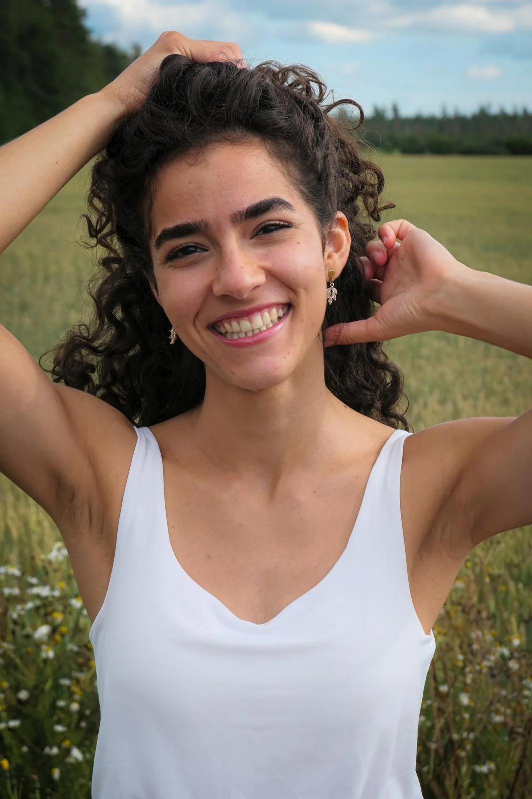 Lächelnde junge Frau mit dunklen, lockigen Haaren, trägt weiße Top, steht in einem Feld mit Blumen, Blick in die Kamera, im Freien bei bewölktem Himmel.