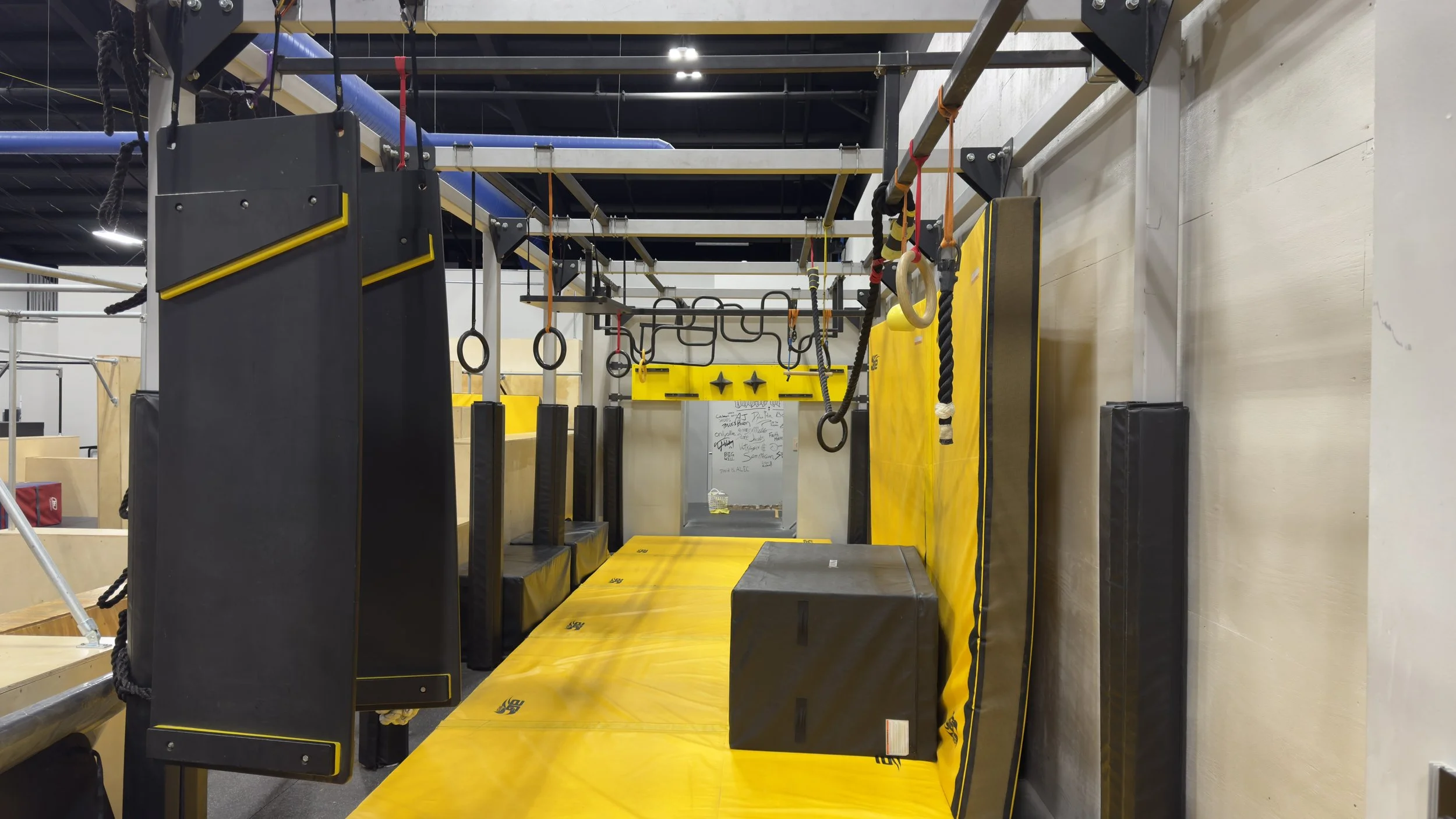 Indoor ninja warrior obstacle course with rings, ropes, walls, and padded yellow flooring.