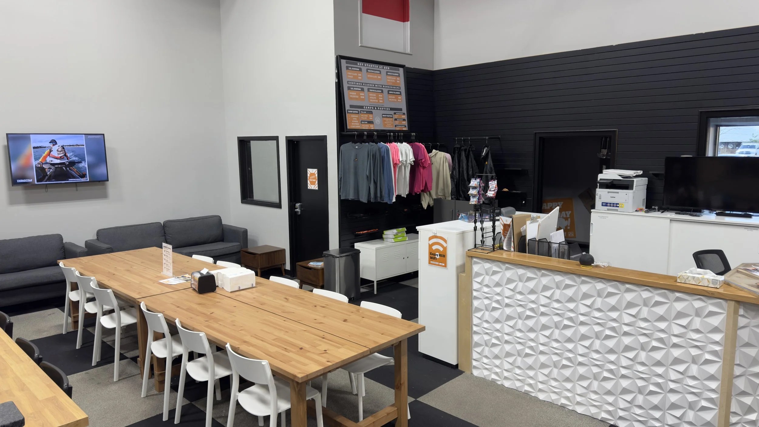 Indoor space with a large wooden table surrounded by white chairs, gray couches against the wall, a black wall with merchandise and signage, a reception desk with a geometric pattern front, and a television mounted on the wall displaying a person in 